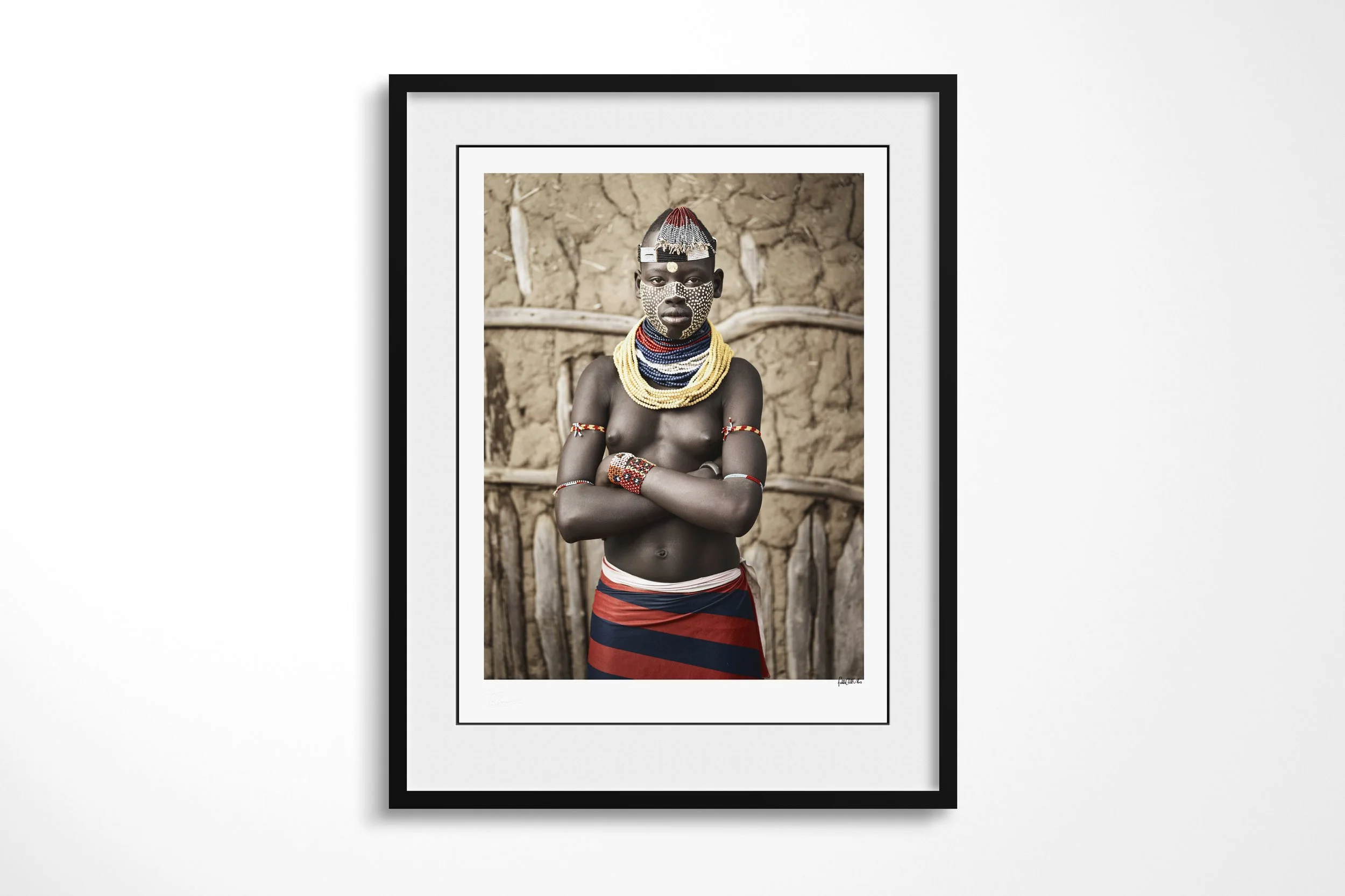 Framed presentation of Tena in a black frame and white mount, showing a Kara woman with patterned facial paint, layered beadwork and folded arms before an earthen wall.