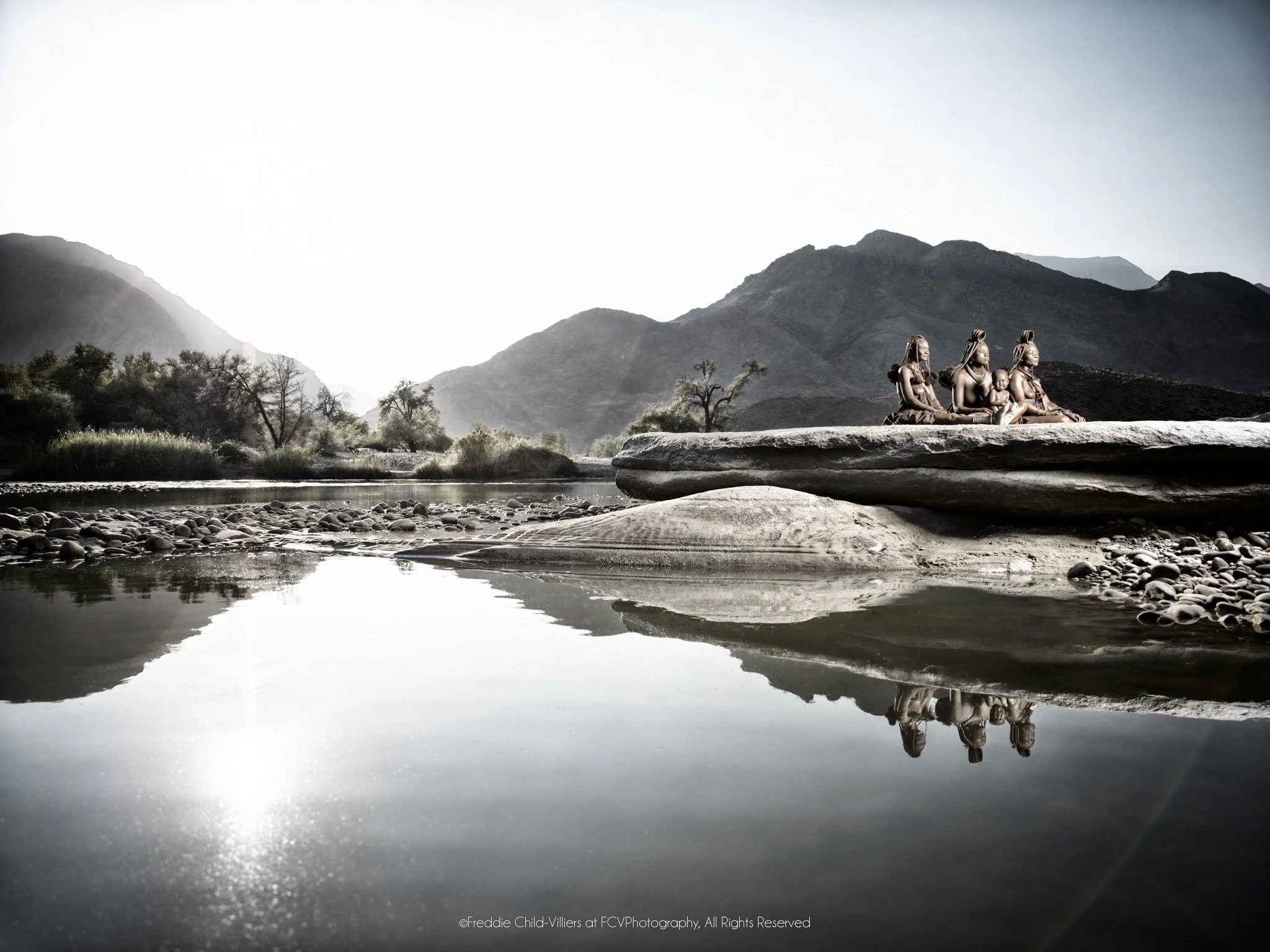 Mbendura (left), Tjikuria and child (middle), Mutume (right), a Himba family gathered on a broad rock beside the Kunene River with their reflections in the water.