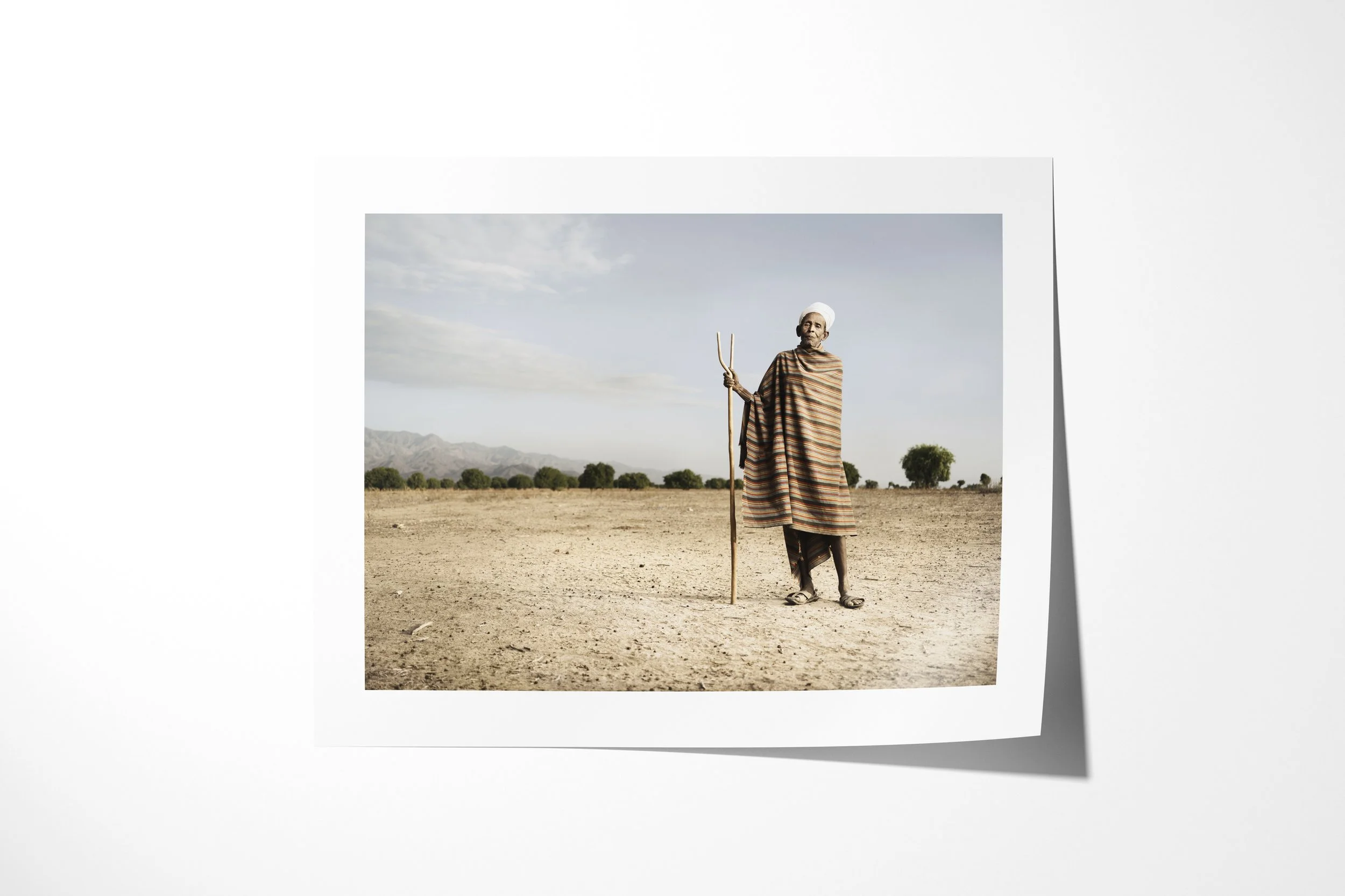 Fine art paper print of Aregodo, showing an older Arbore man in a striped wrap and white head cloth holding a forked staff on a dry plain.