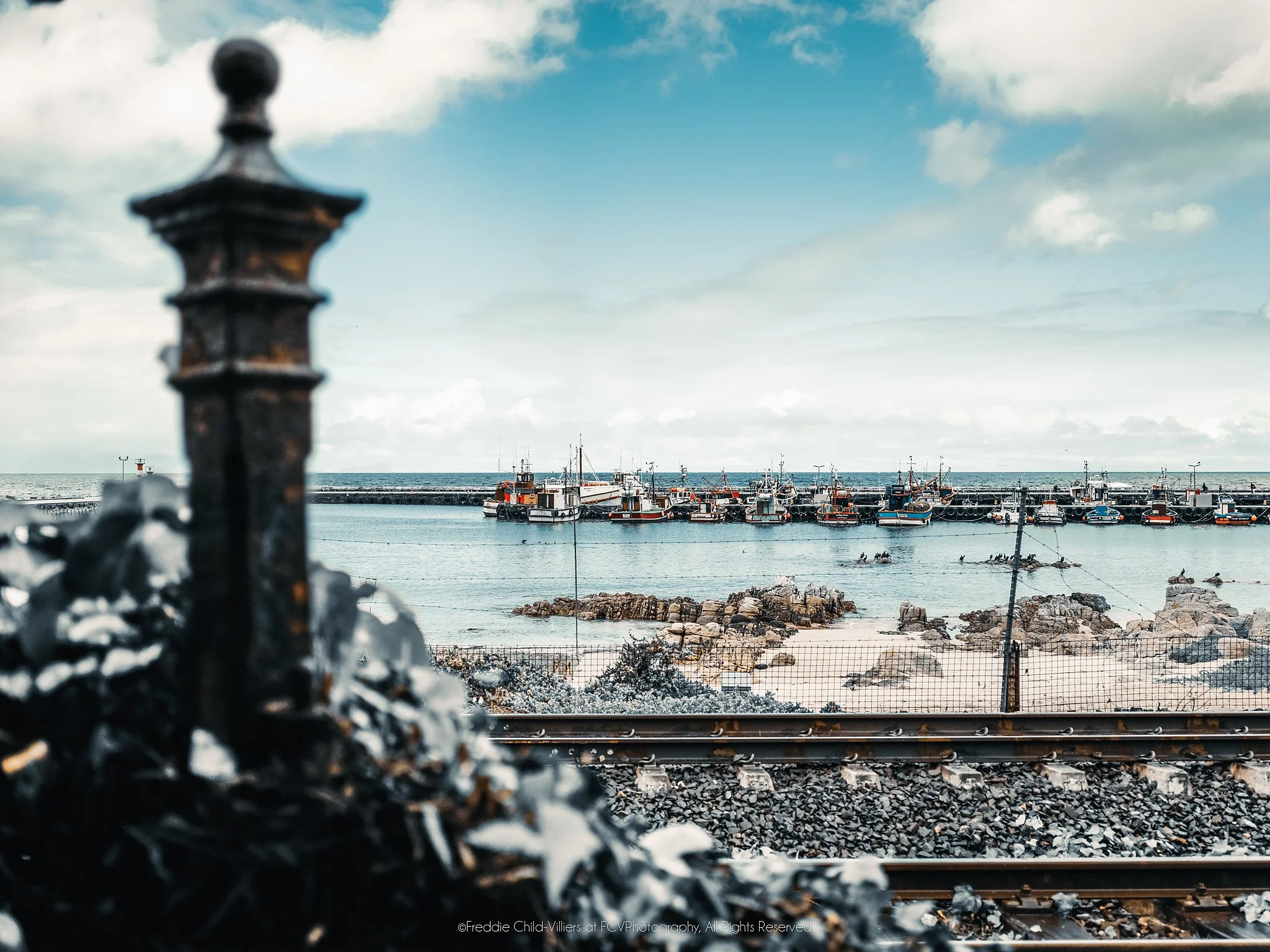 Fishing boats moored across a calm harbour beyond a rocky shore and railway tracks, with a blurred iron post in front.