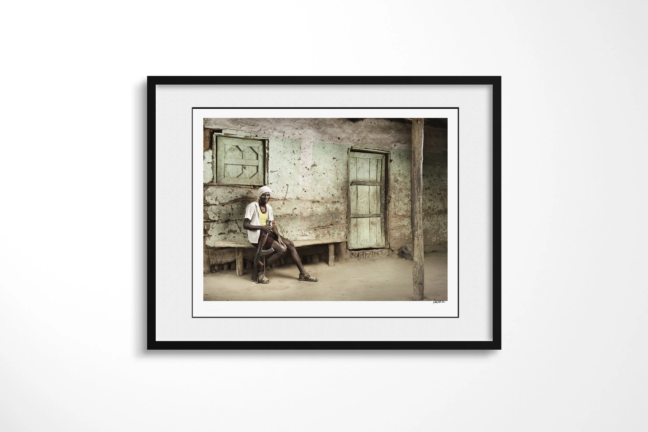 Framed presentation of Arshal in a black frame and white mount, showing an Arbore man seated with a rifle outside a weathered house with green shutters.