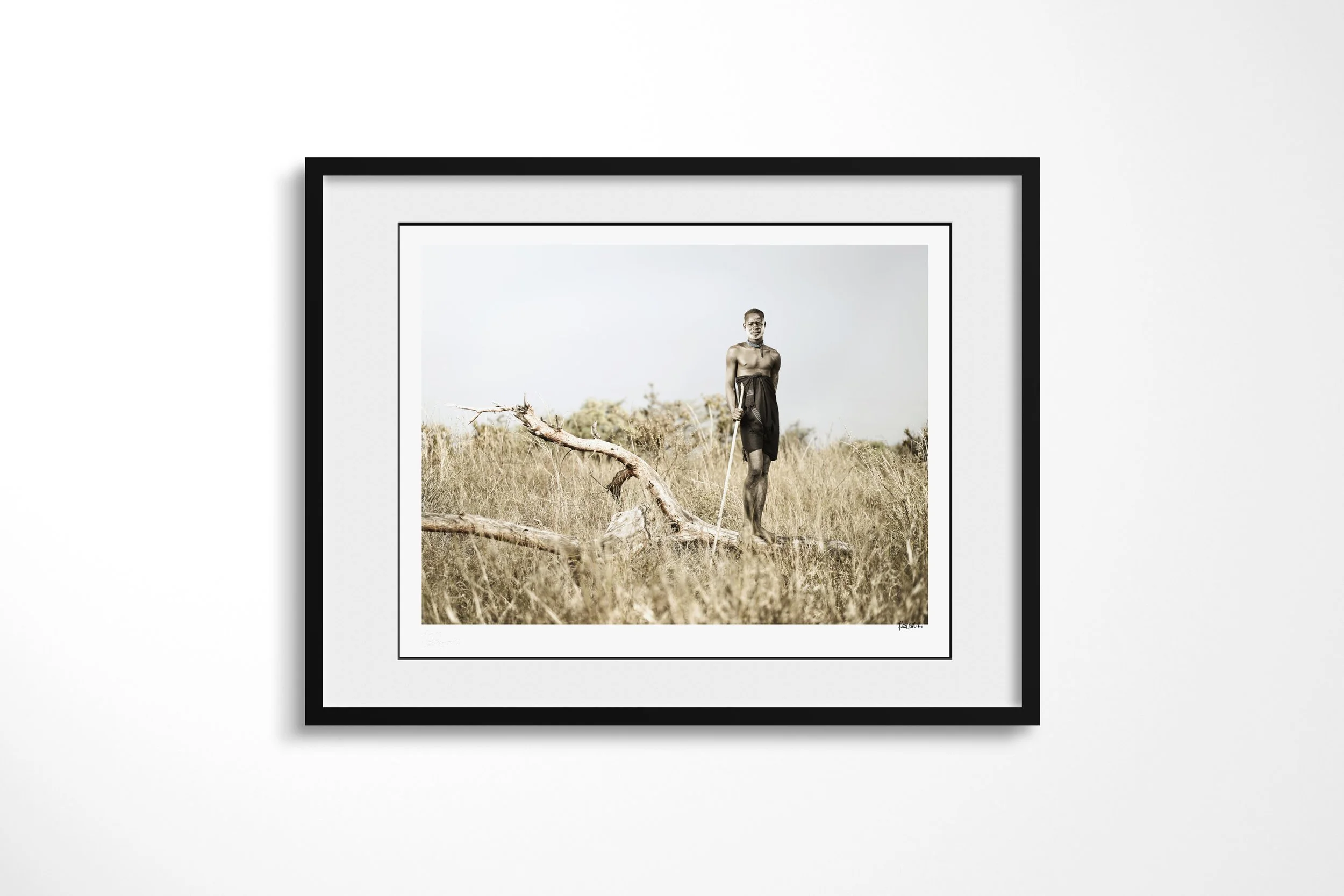 Framed presentation of Morkoro in a black frame and white mount, showing a Mursi man in a dark wrap standing with a staff beside a fallen branch in dry grassland.