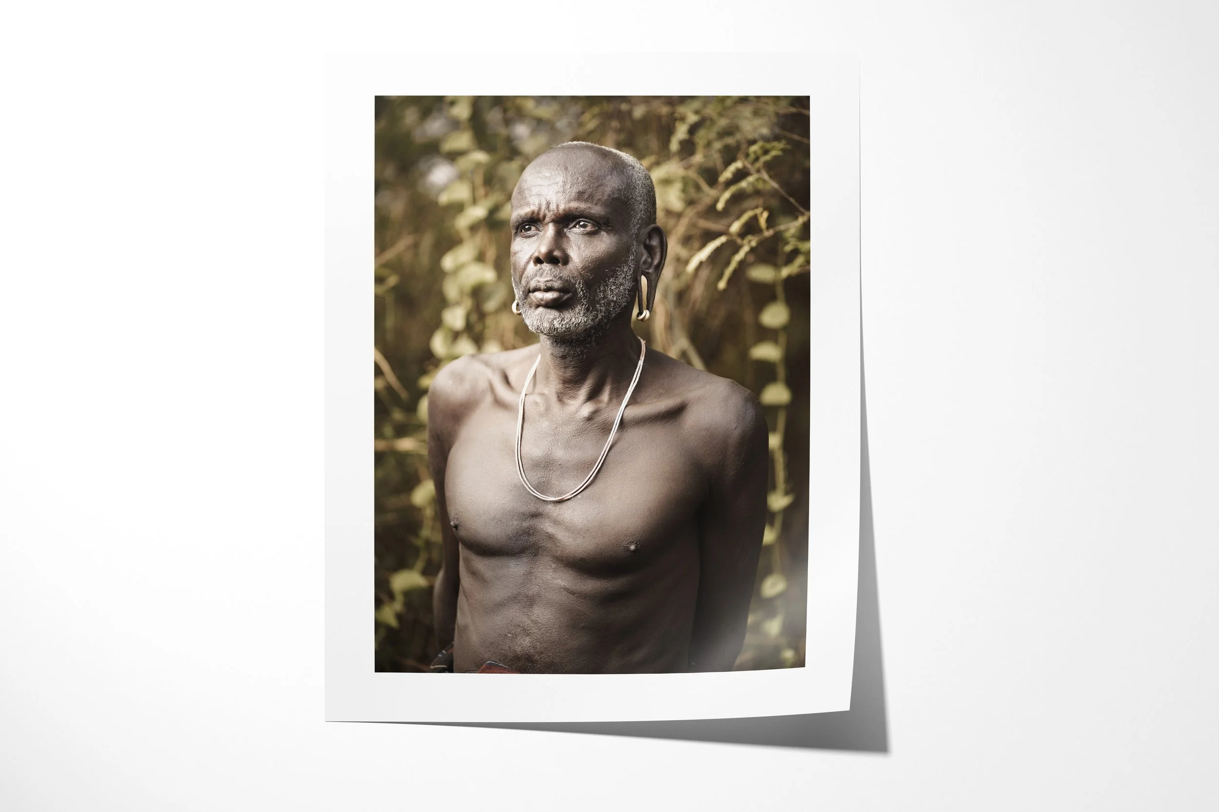 Fine art paper print of Bardhoro, showing an older Mursi man shown bare-chested in a close portrait against soft woodland.