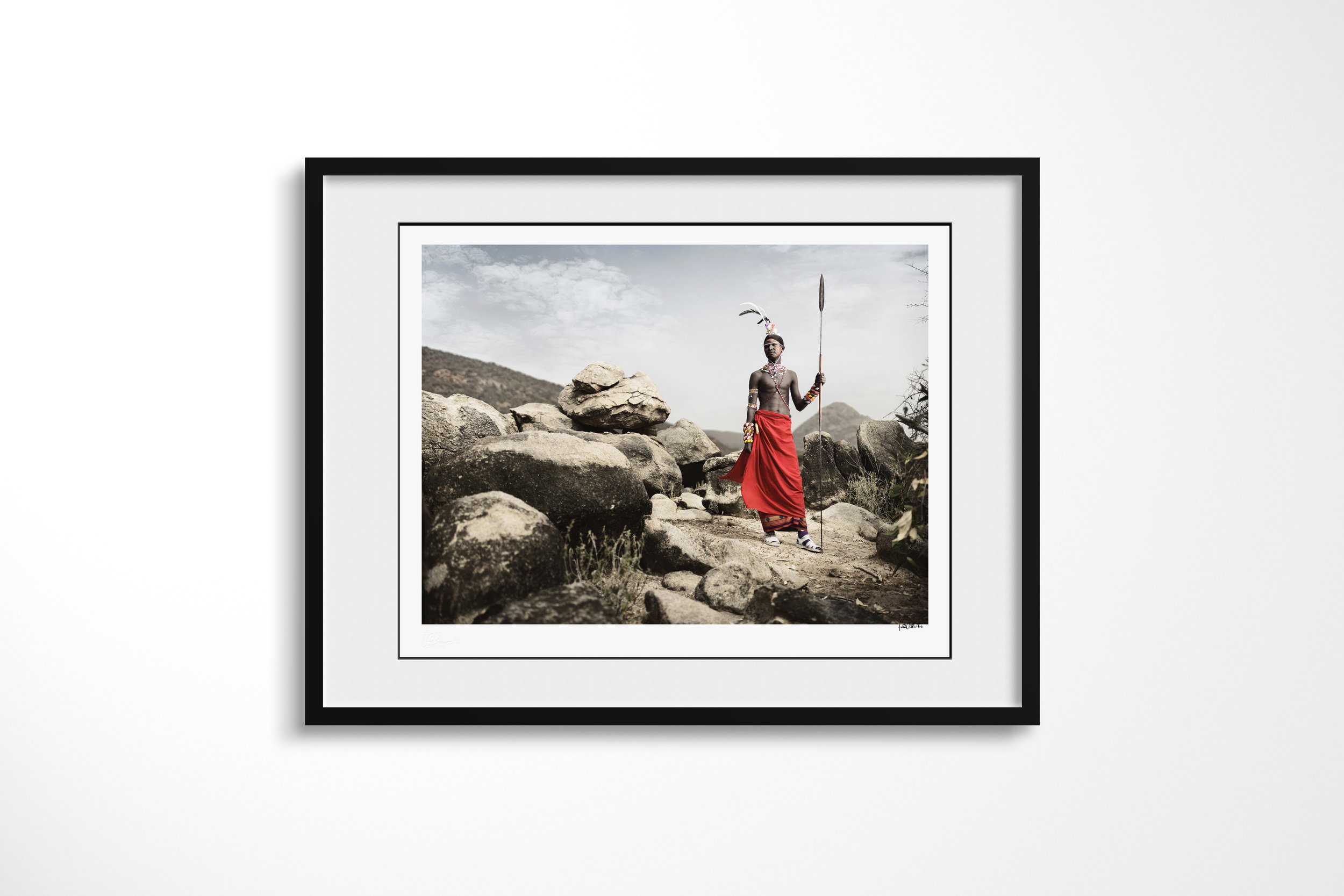 Framed presentation of Lempati in a black frame and white mount, showing a Samburu warrior in a red wrap holding a spear among large rocks and mountain scrub.