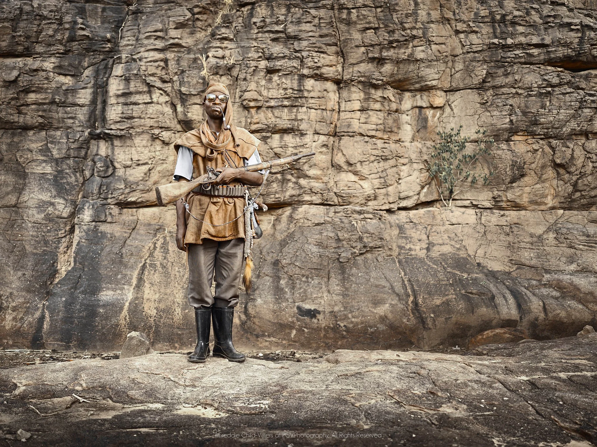 Diongo standing with a rifle, ammunition belt and boots before a rock wall.