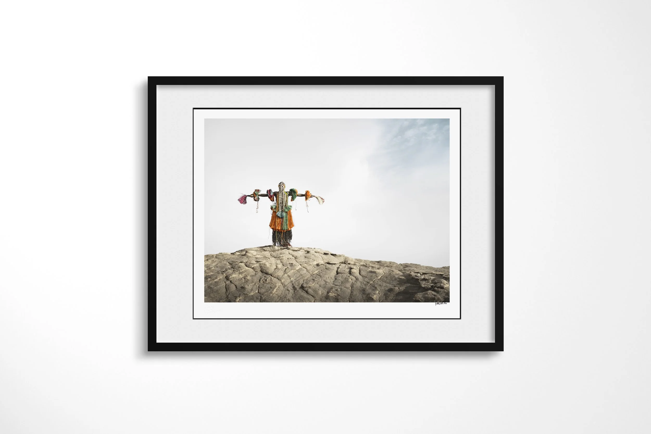 Framed presentation of Legada Iyayi (Mask) in a black frame and white mount, showing a Dogon bird mask with outstretched arms standing on a rock ridge.