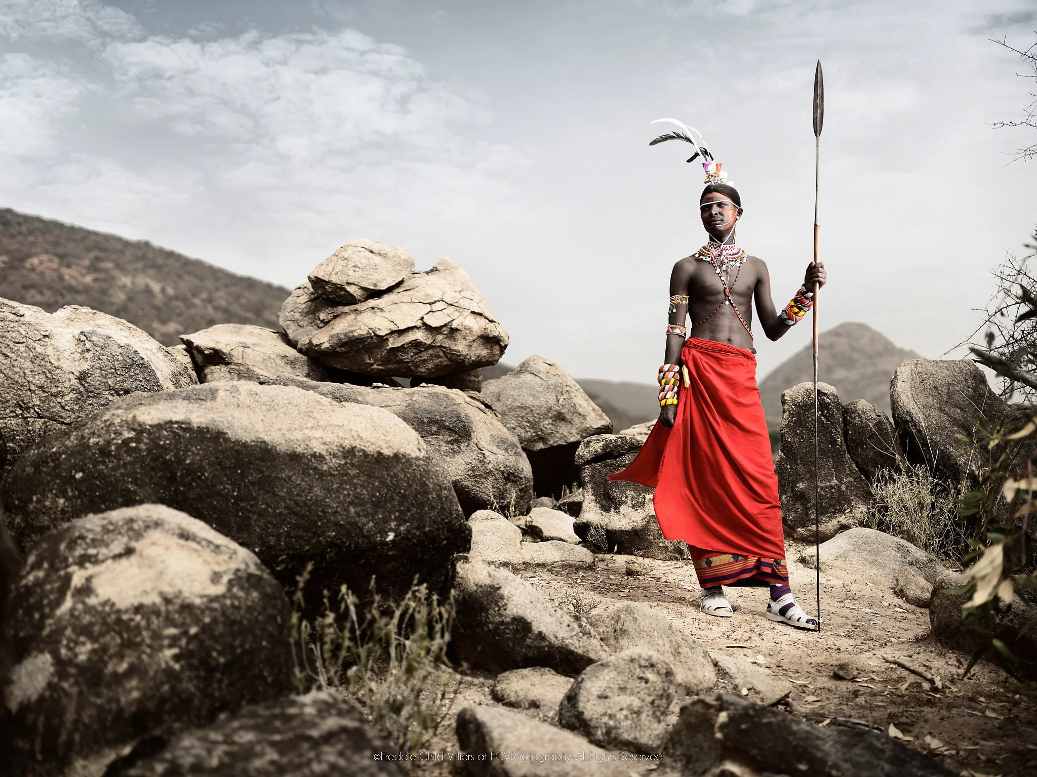 Lempati, a Samburu warrior in a red wrap holding a spear among large rocks and mountain scrub.