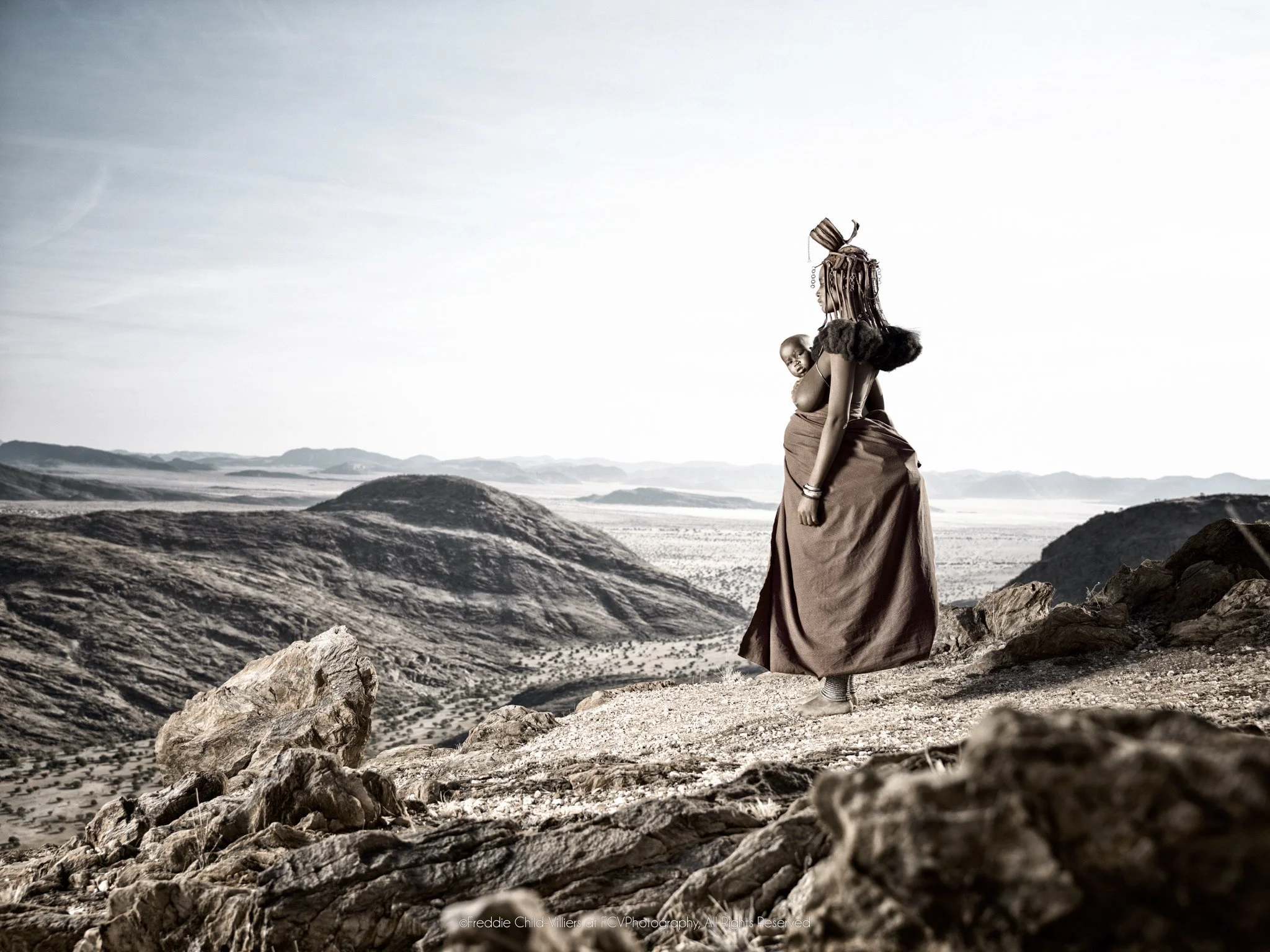Venongore carrying her child on her back while looking across the rocky Marienfluss.