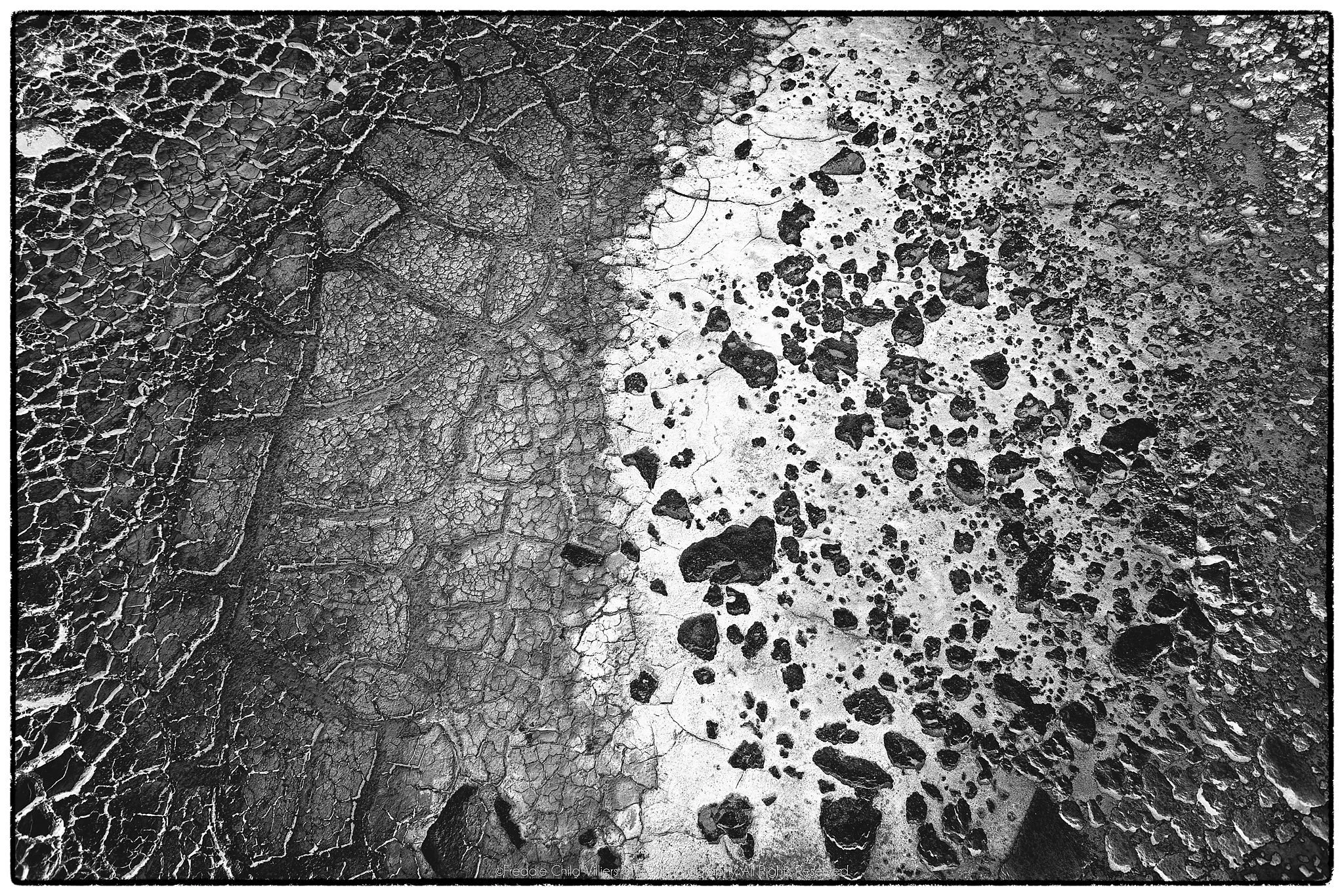 Cracked earth in black and white, divided by a broad pale curving band scattered with dark stones.