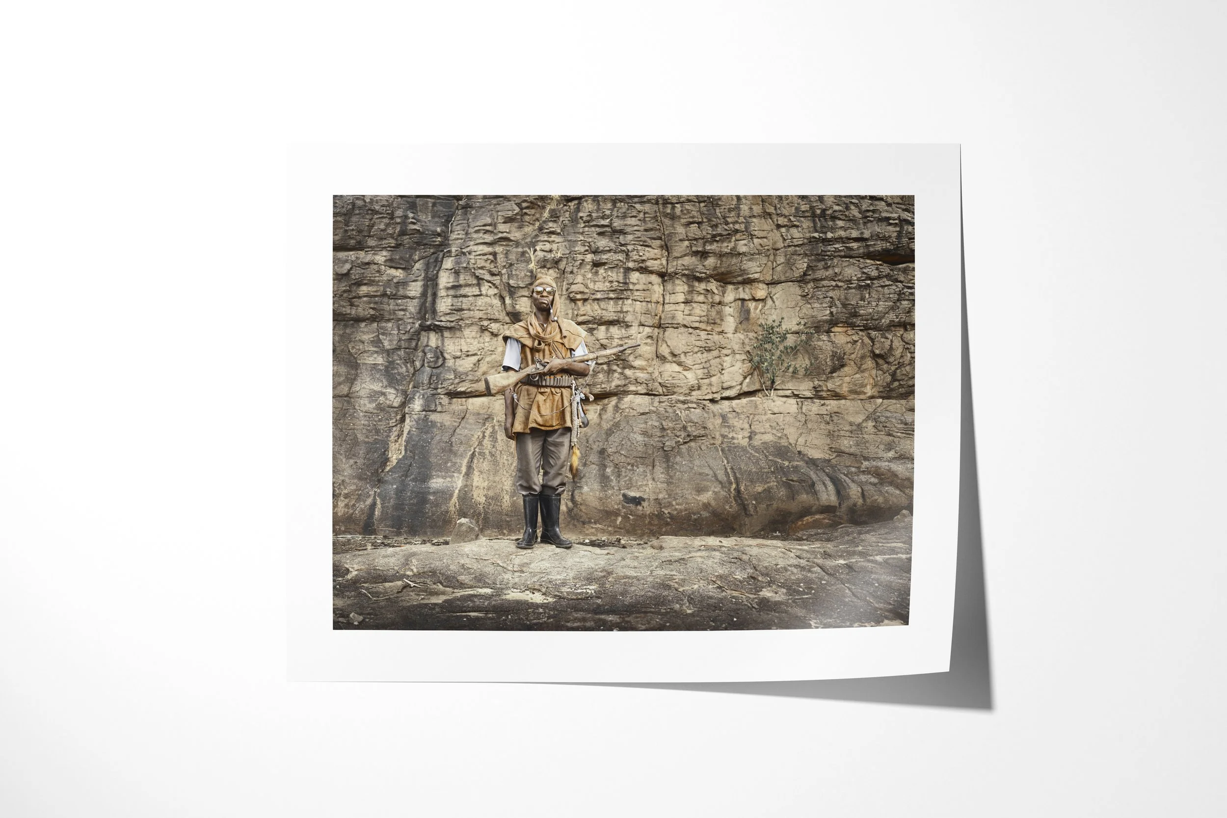 Fine art paper print of Diongo, showing Diongo standing with a rifle, ammunition belt and boots before a rock wall.