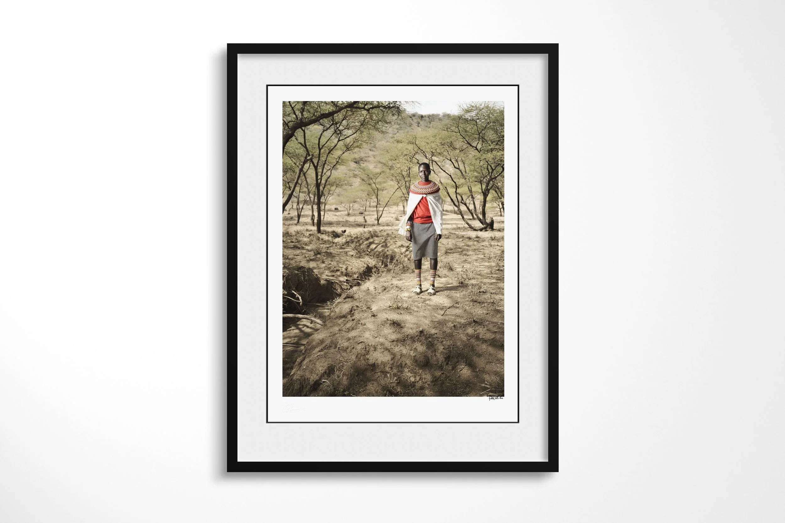 Framed presentation of Noswuray in a black frame and white mount, showing a Samburu woman wearing a white shawl and broad bead collar on a path through open woodland.