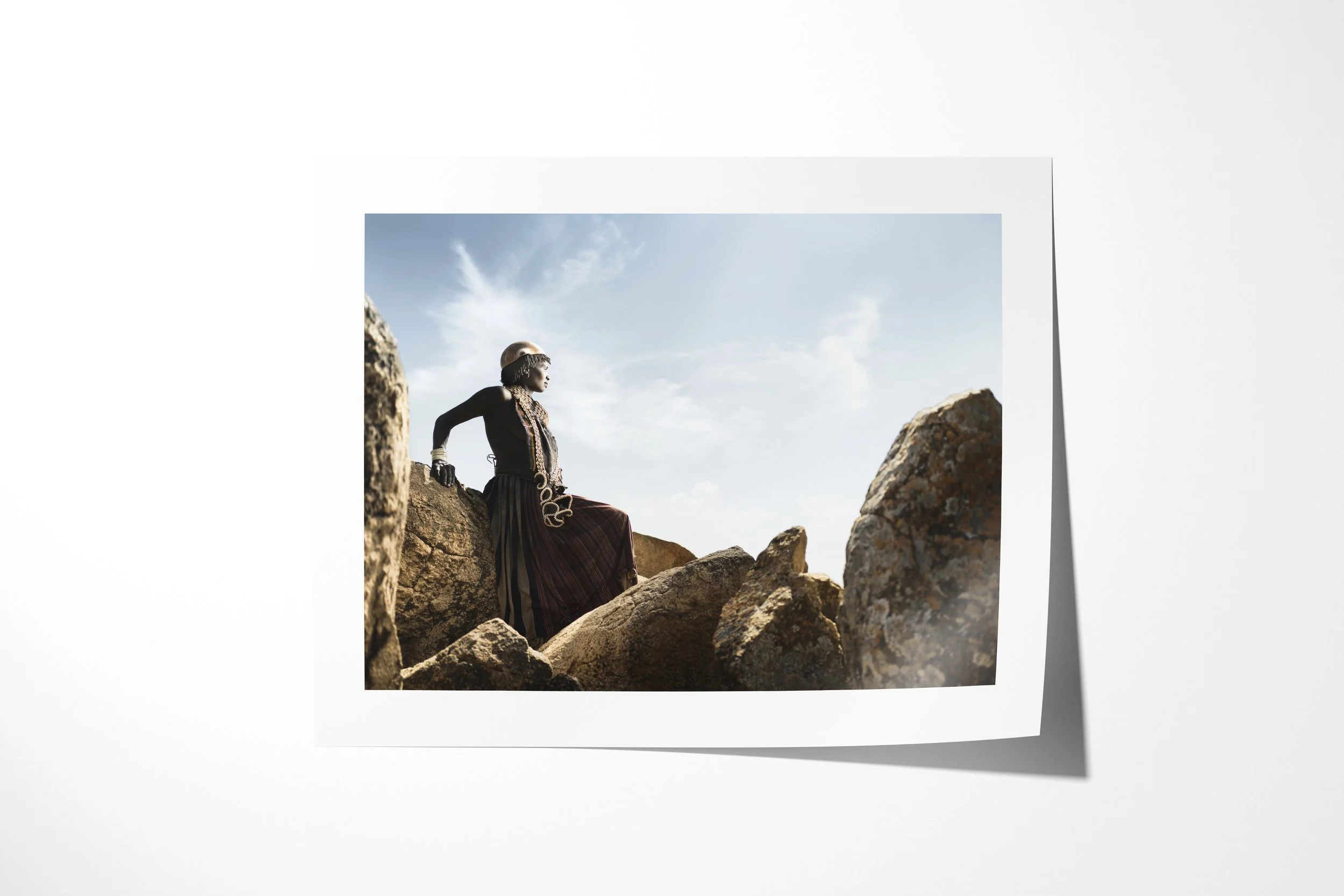 Fine art paper print of Koiga, showing a Banna woman in a long dark dress sitting among large sunlit rocks beneath an open sky.