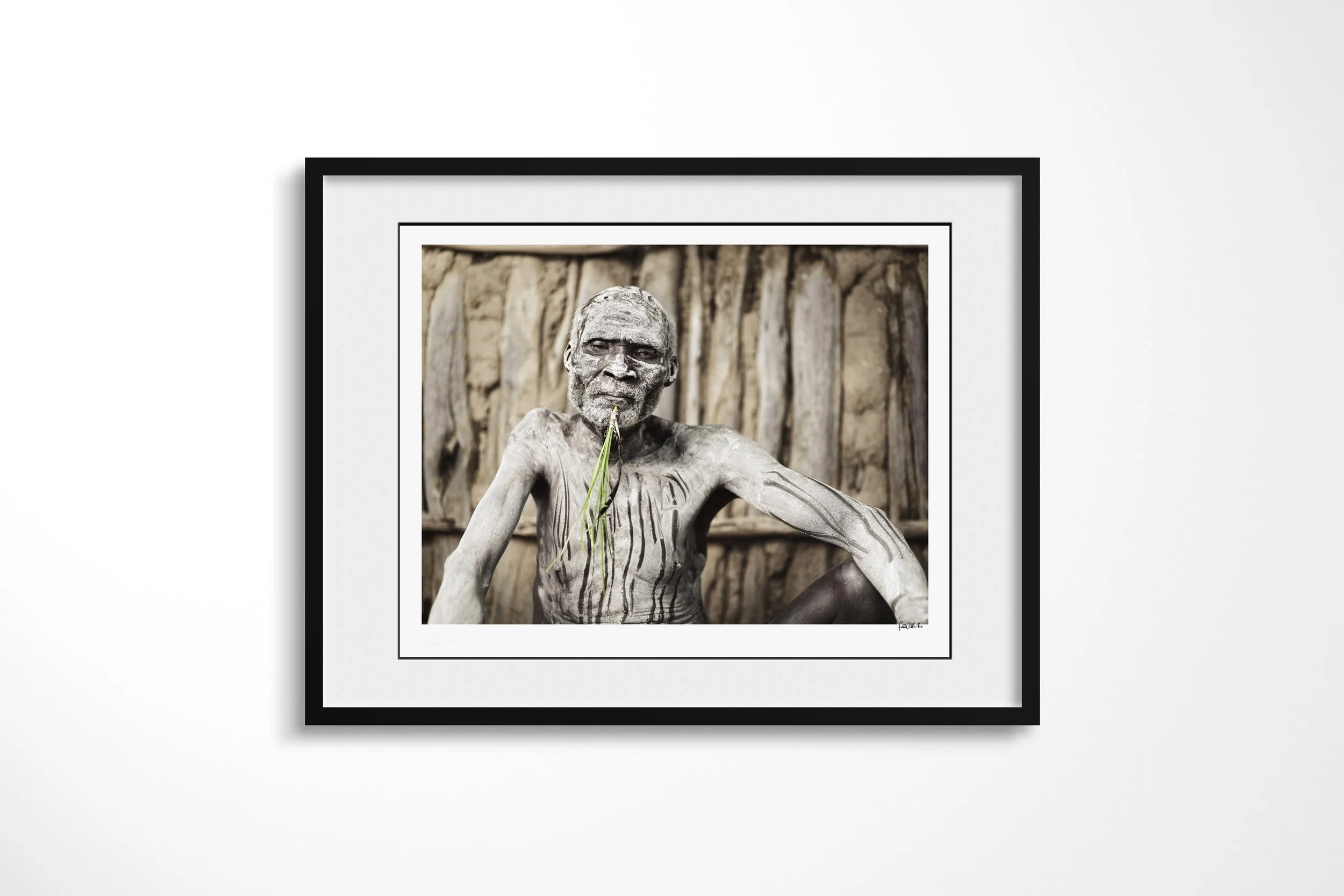 Framed presentation of Bonko in a black frame and white mount, showing an older Kara man with pale body paint and dark painted lines seated against a wooden wall.