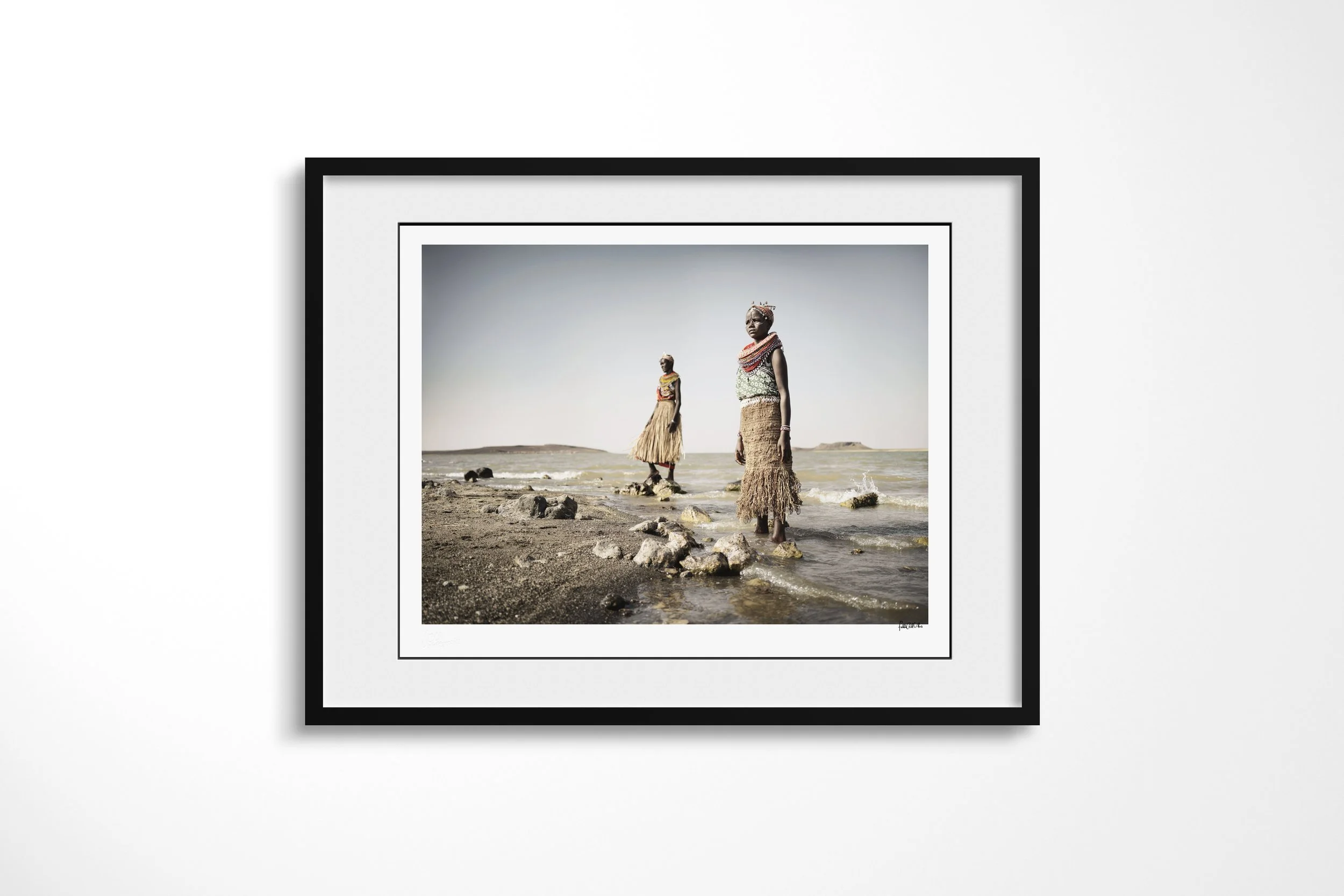Framed presentation of Lawra & Nadirie in a black frame and white mount, showing two El Molo women in fibre garments standing on the rocky shore of Lake Turkana.