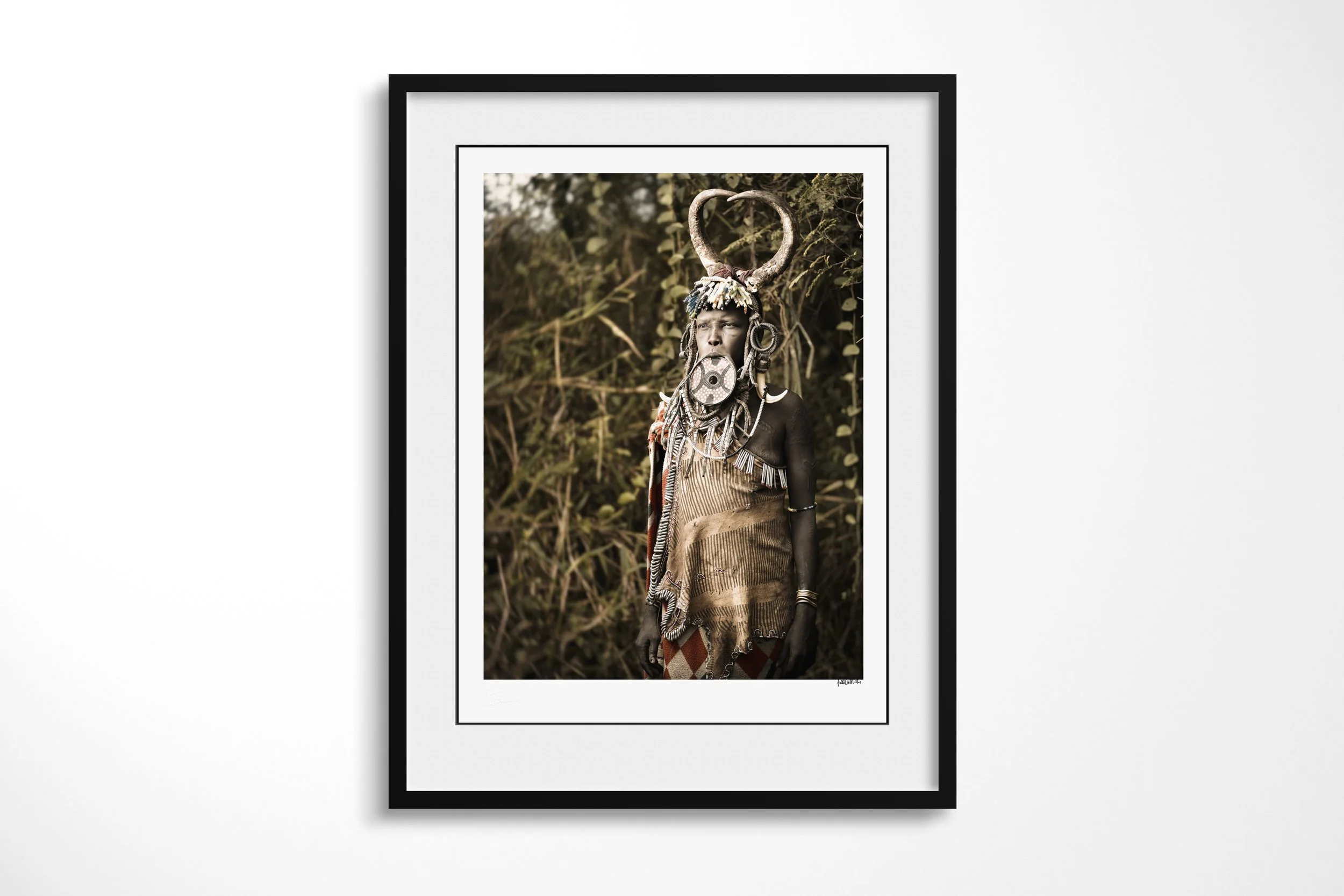 Framed presentation of Madhawori in a black frame and white mount, showing a Mursi woman wearing a painted lip plate, beadwork and a circular horn headdress.
