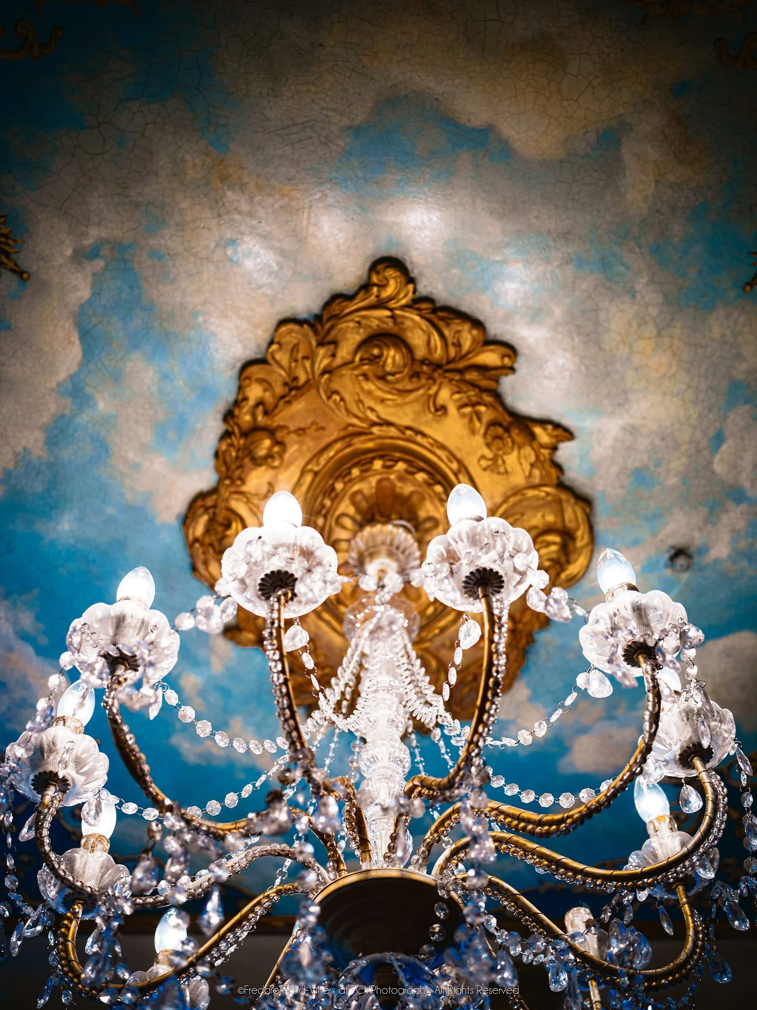 Chandelier looks upward through crystal drops and curved arms to a gilded ceiling rose set against cracked, timeworn blue plaster and reflected light.