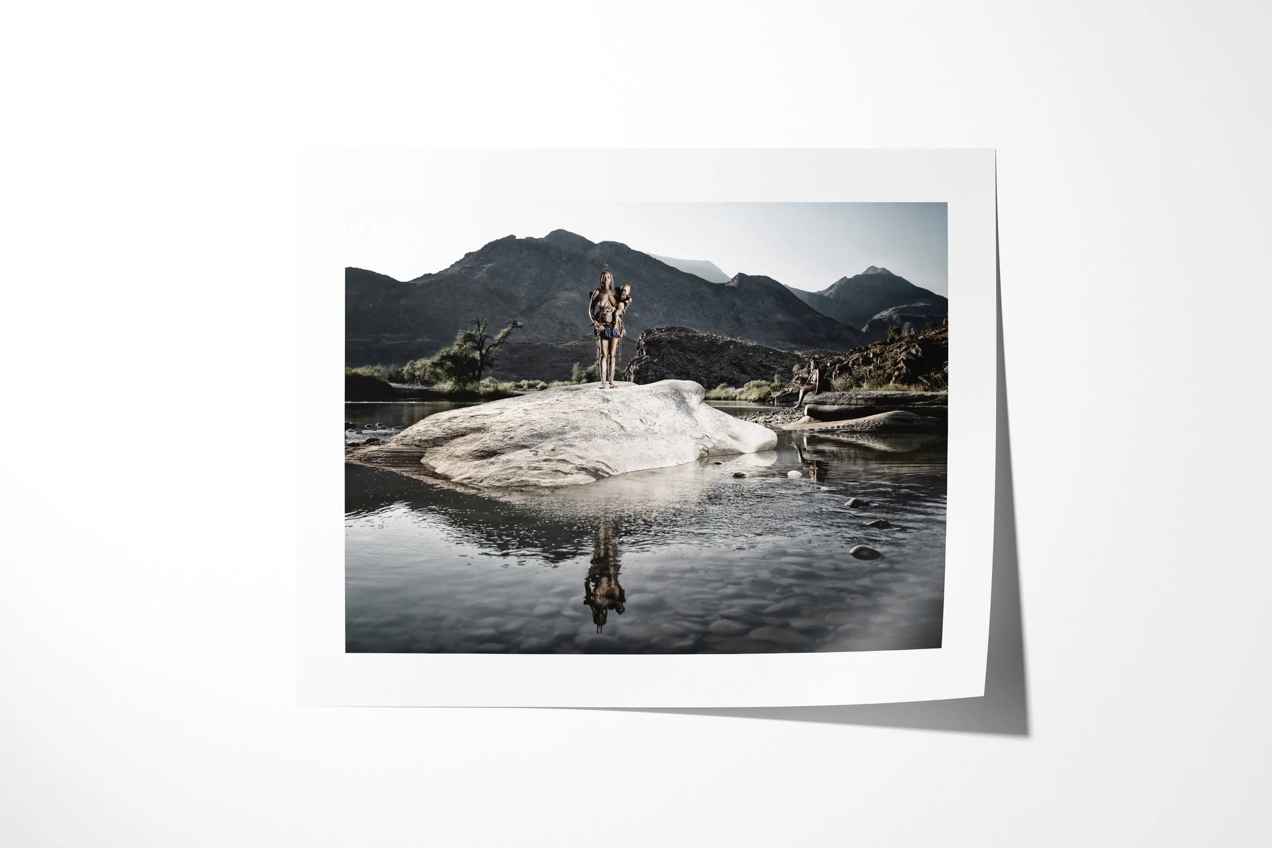 Fine art paper print of Tunae (foreground), Katafa (background), showing two distant figures standing on a river boulder, reflected in the still water below.