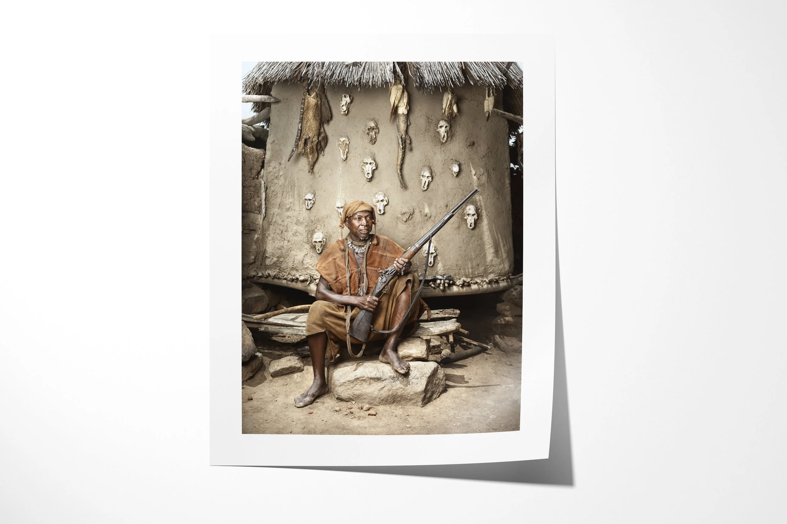 Fine art paper print of Lion, showing Lion seated with a flintlock rifle before an earthen wall set with animal skulls.