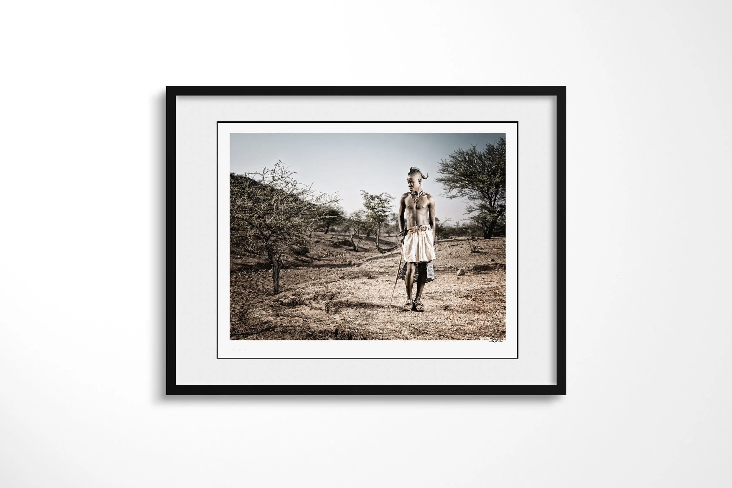 Framed presentation of Hembinda in a black frame and white mount, showing Hembinda holding a staff on a path through sparse dry woodland.