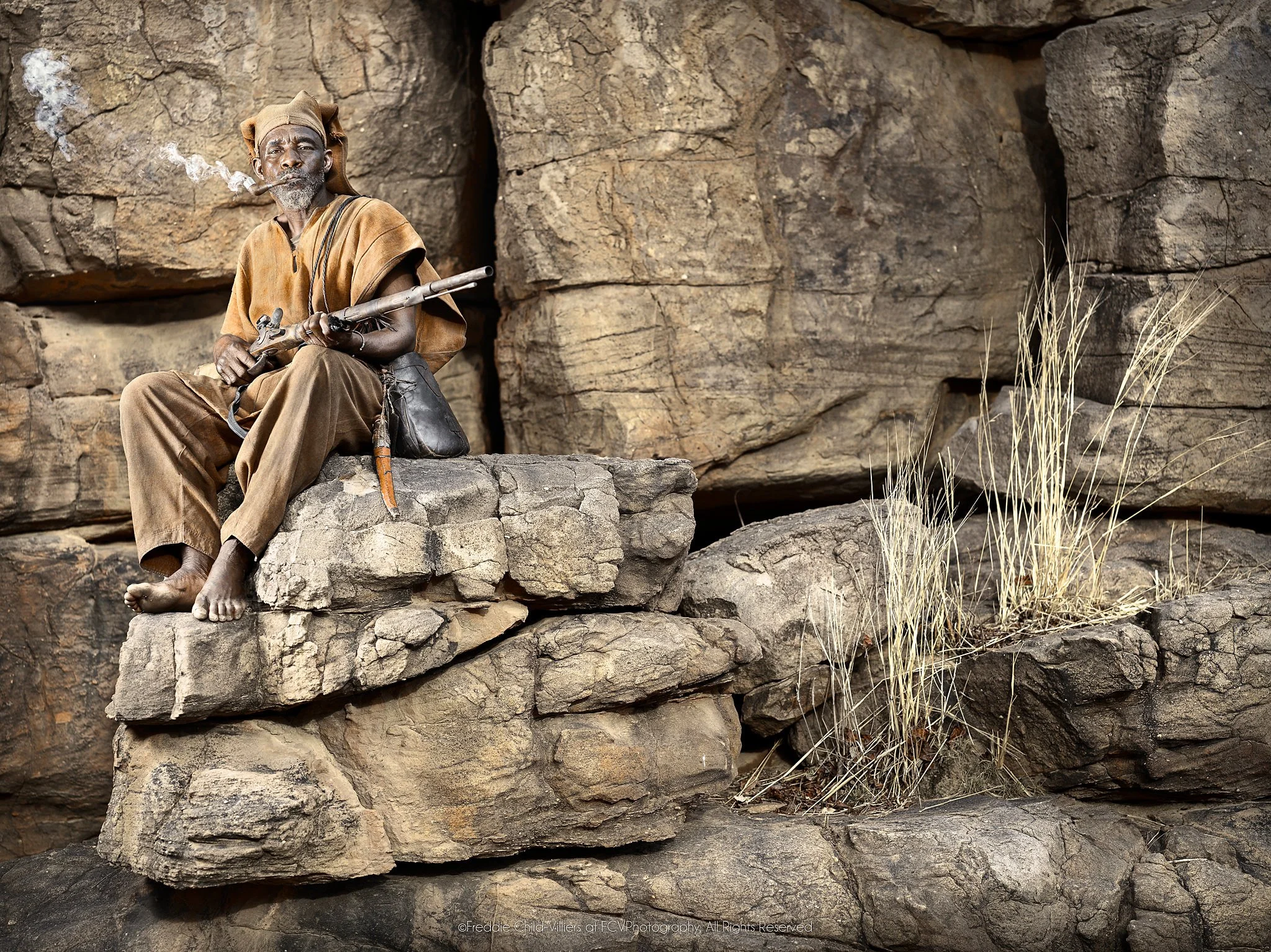 Antandou seated on layered rock with a rifle as pipe smoke rises beside him.
