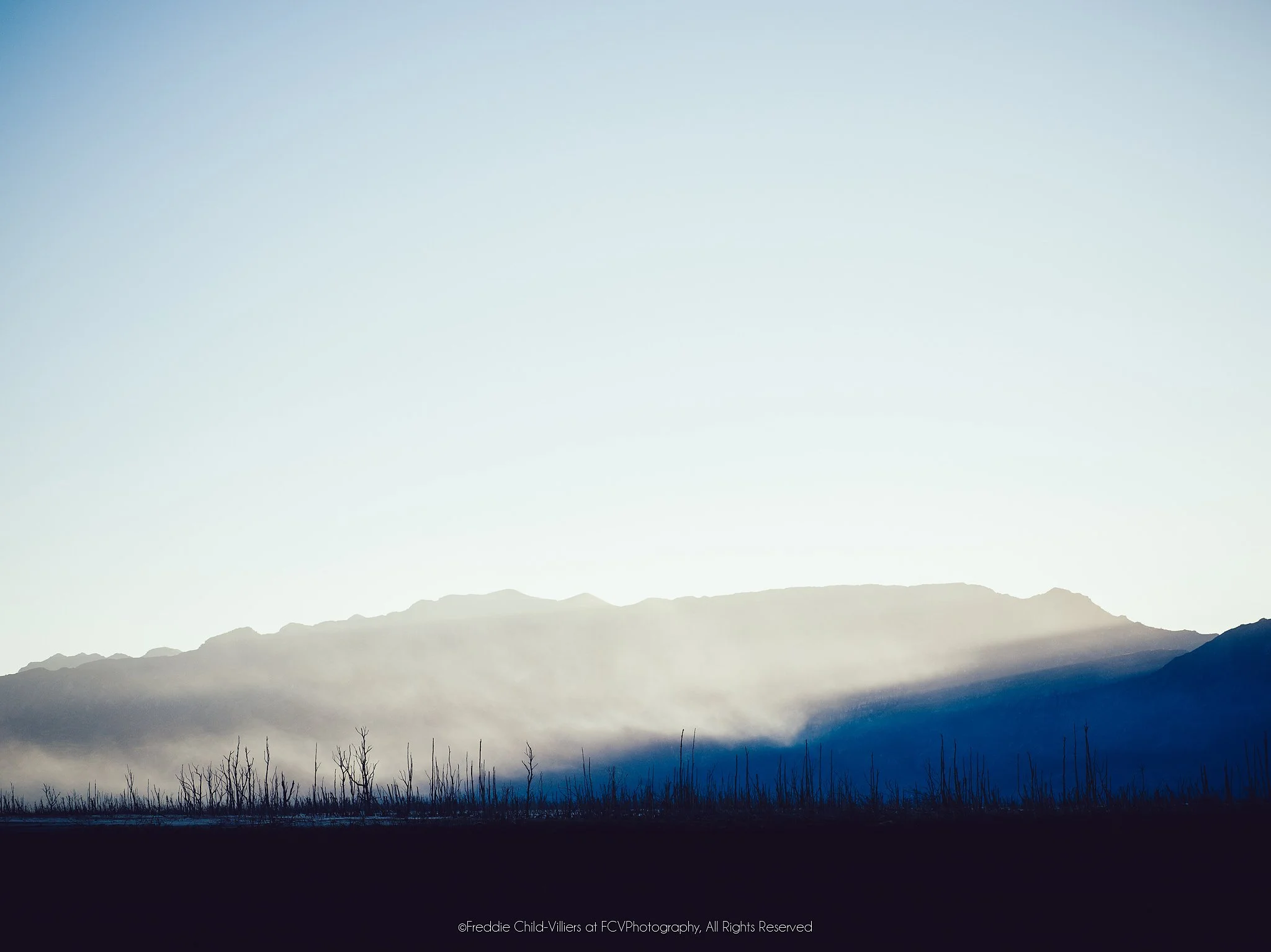 Bare blackened trees below a mountain ridge, with pale mist lit above a deep blue shadow.