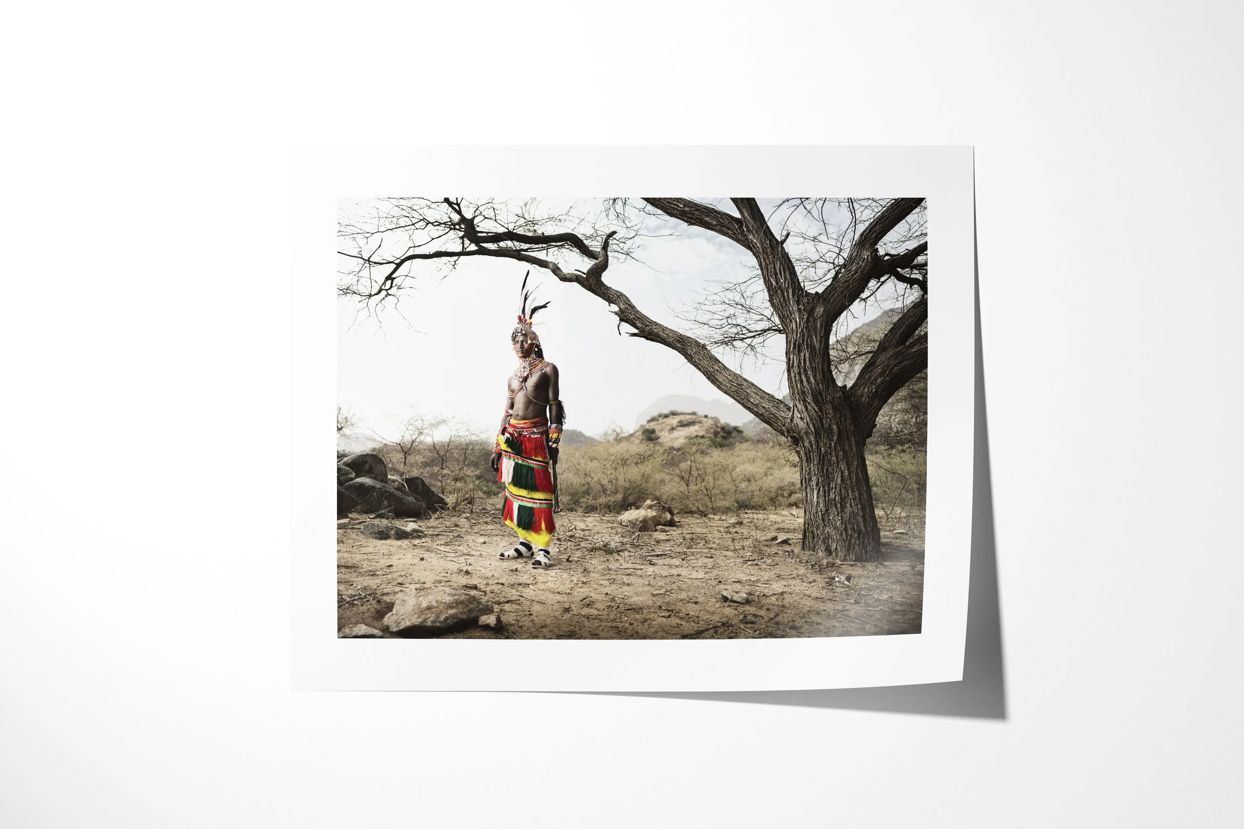Fine art paper print of Lebitili, showing a Samburu warrior in colourful beadwork standing beneath a leafless tree in rocky country.