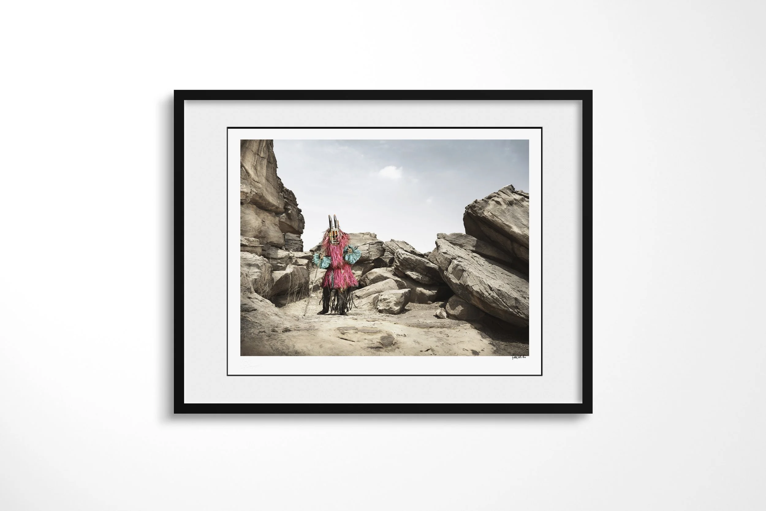 Framed presentation of Walu (Mask) in a black frame and white mount, showing a Dogon masked figure in bright fibre costume standing in a narrow rocky passage.
