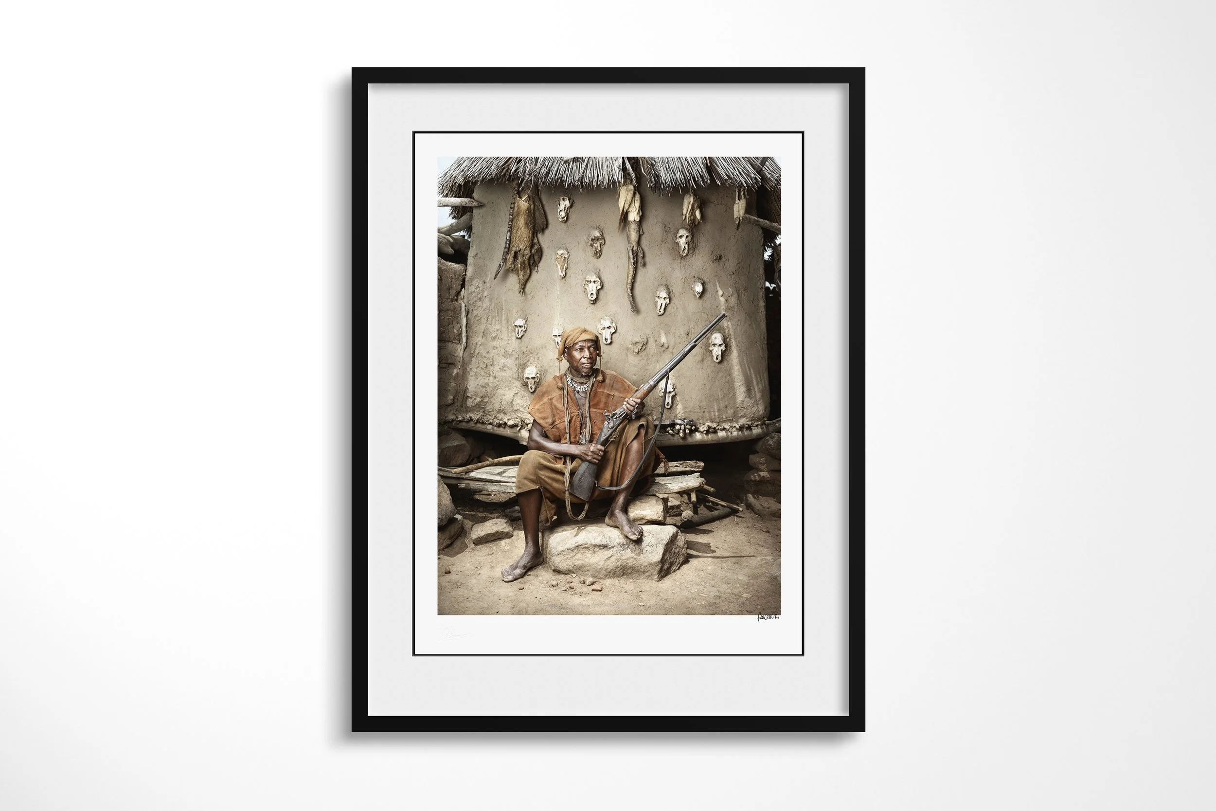 Framed presentation of Lion in a black frame and white mount, showing Lion seated with a flintlock rifle before an earthen wall set with animal skulls.