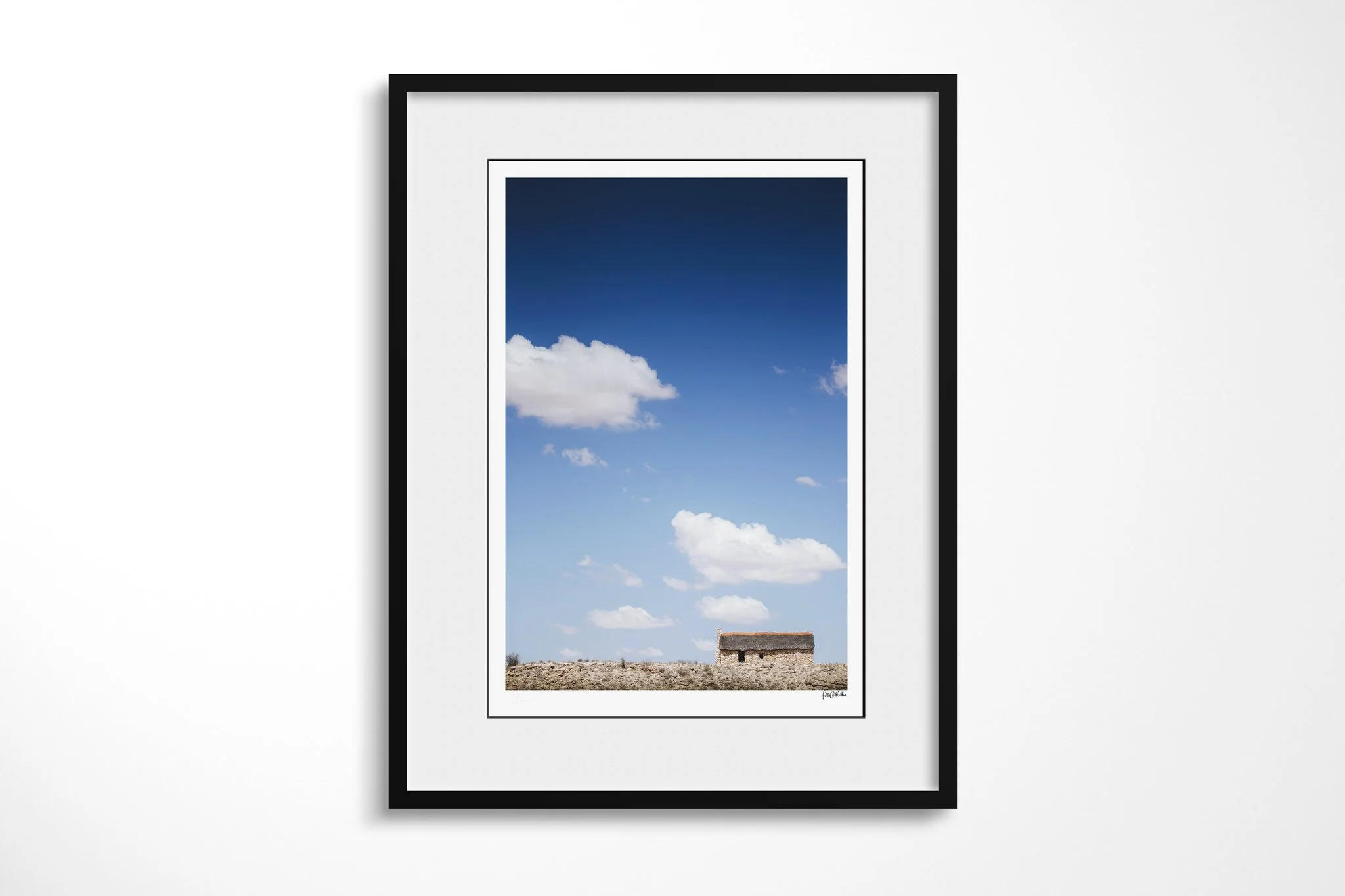 Framed fine art presentation of House On The Hill in a black frame with a white mount, depicting a small thatched house isolated on a barren ridge beneath a vast blue sky and scattered white clouds.