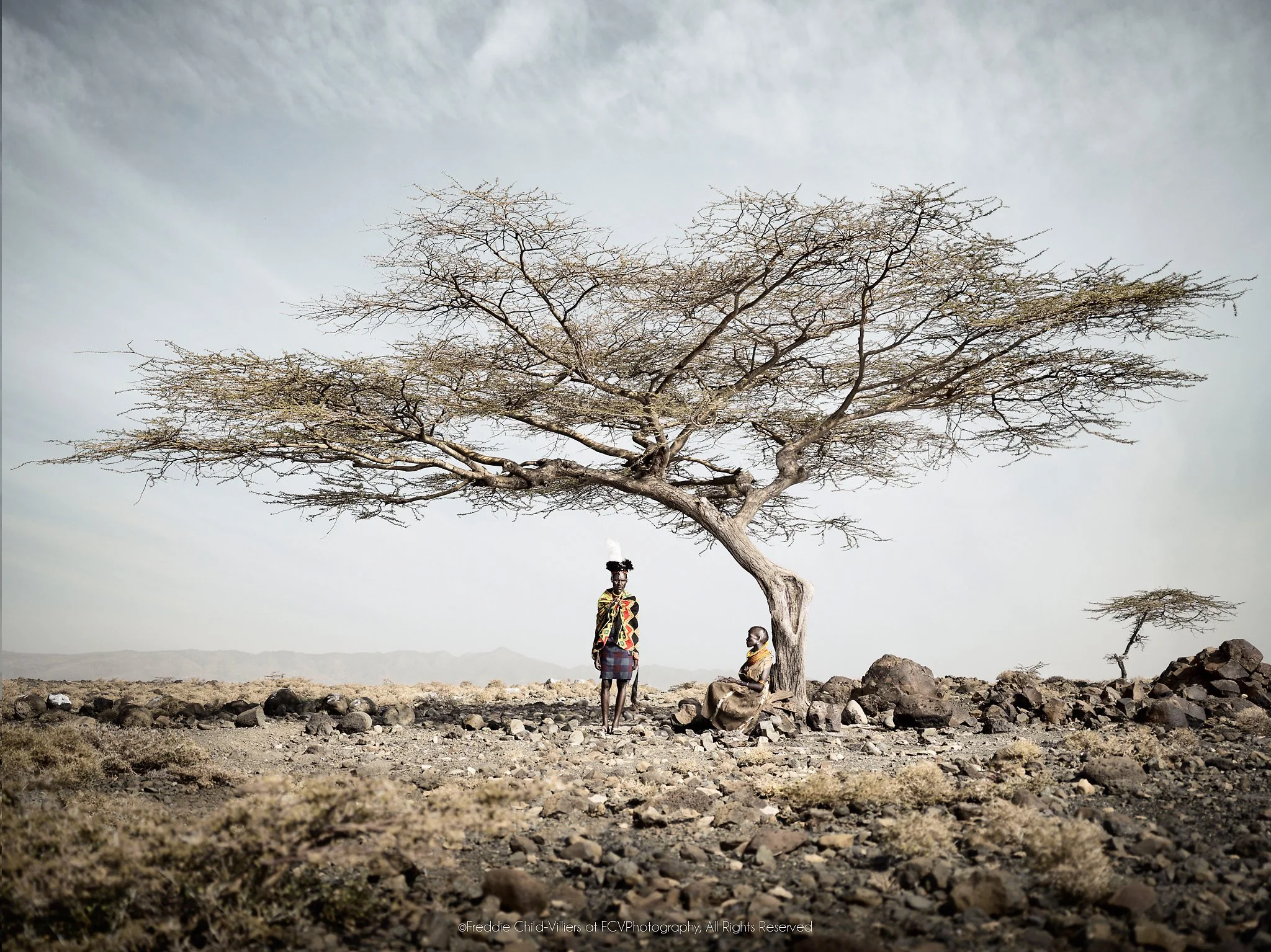 Ayapan and Lotilan standing beneath a broad acacia in a stony Turkana landscape.