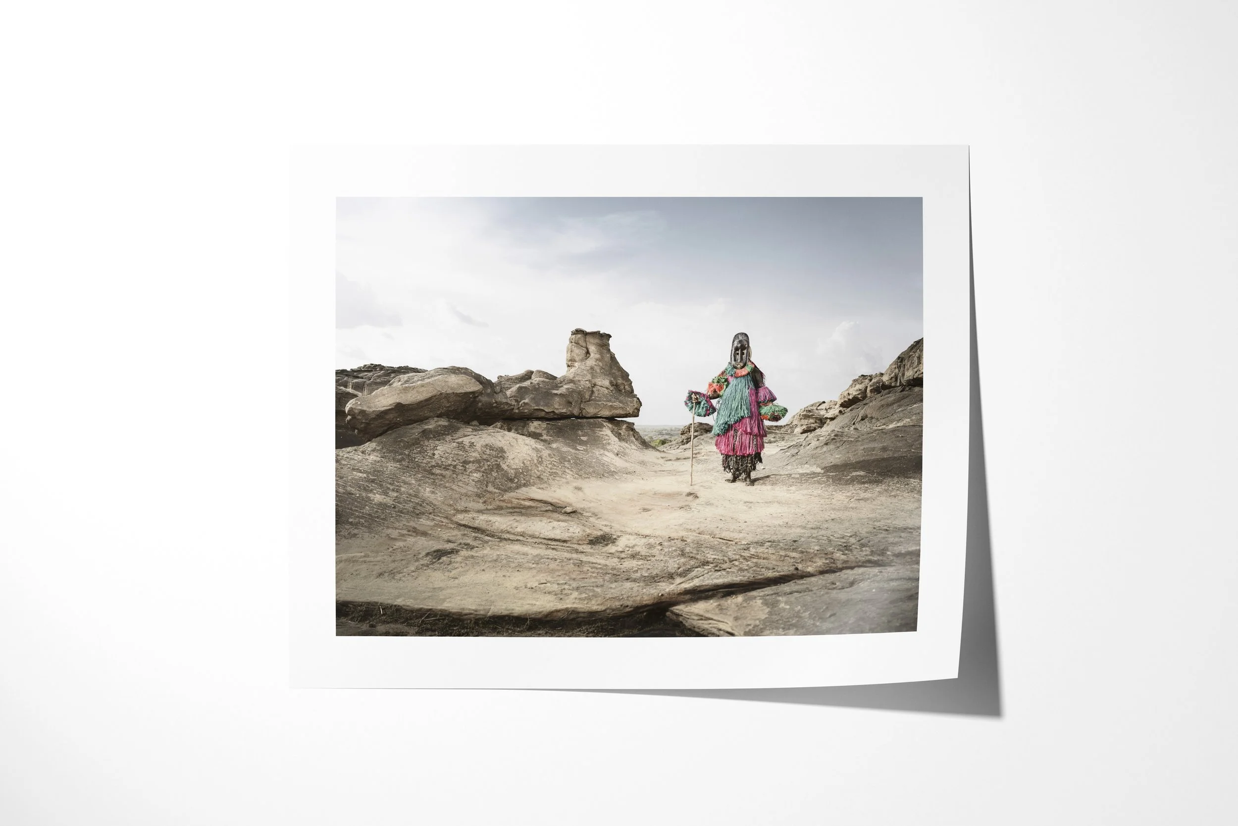 Fine art paper print of Dana (Mask), showing a Dogon hunter mask walking across open rock beneath a pale sky.