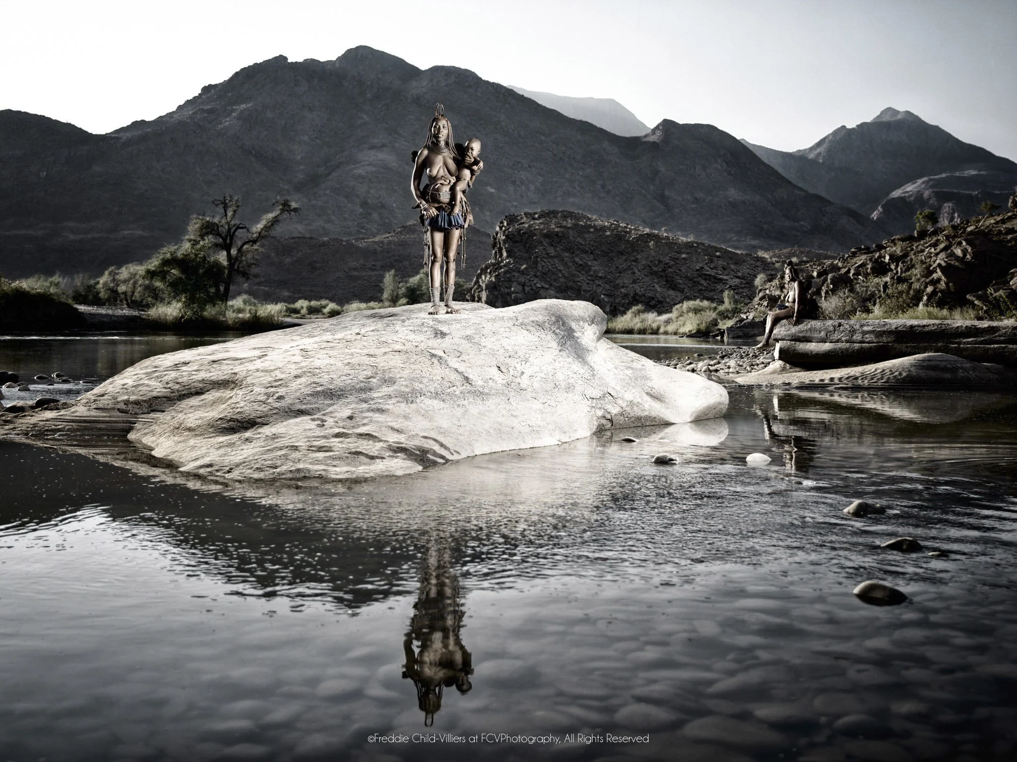 Tunae (foreground), Katafa (background), two distant figures standing on a river boulder, reflected in the still water below.