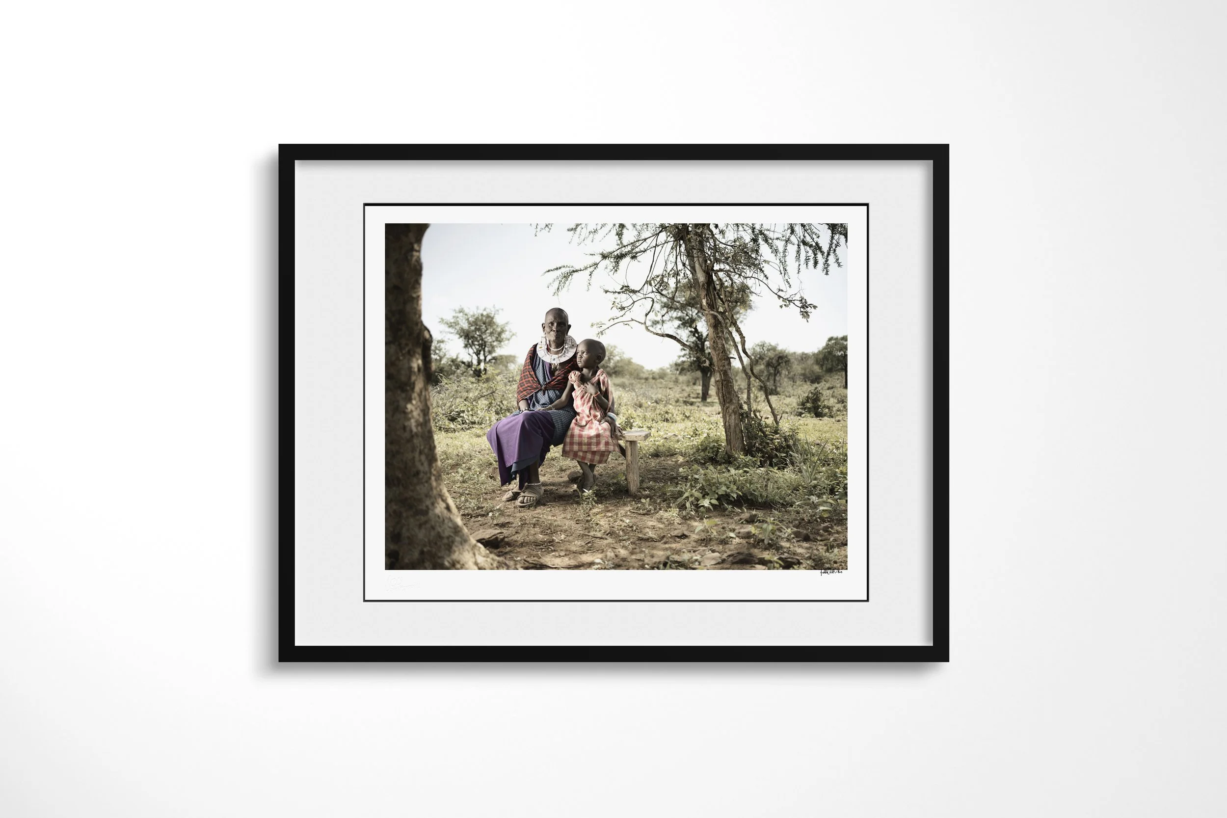 Framed presentation of Naleku & Elias in a black frame and white mount, showing Naleku seated beneath trees with her young grandson Elias standing beside her.