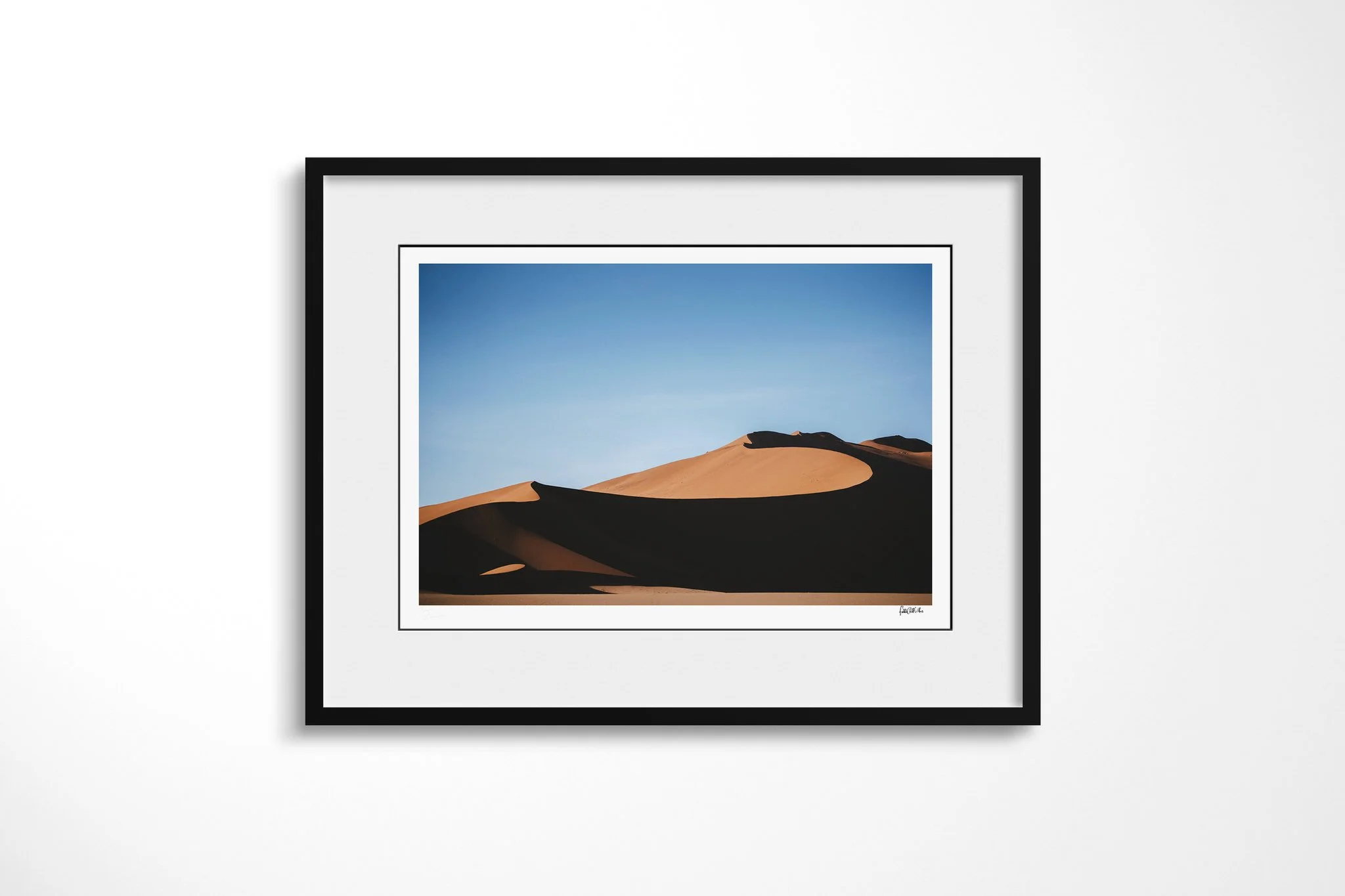 Framed presentation of Surpent in a black frame and white mount, showing sunlit orange desert dunes divided by deep sinuous shadows beneath a clear blue sky.