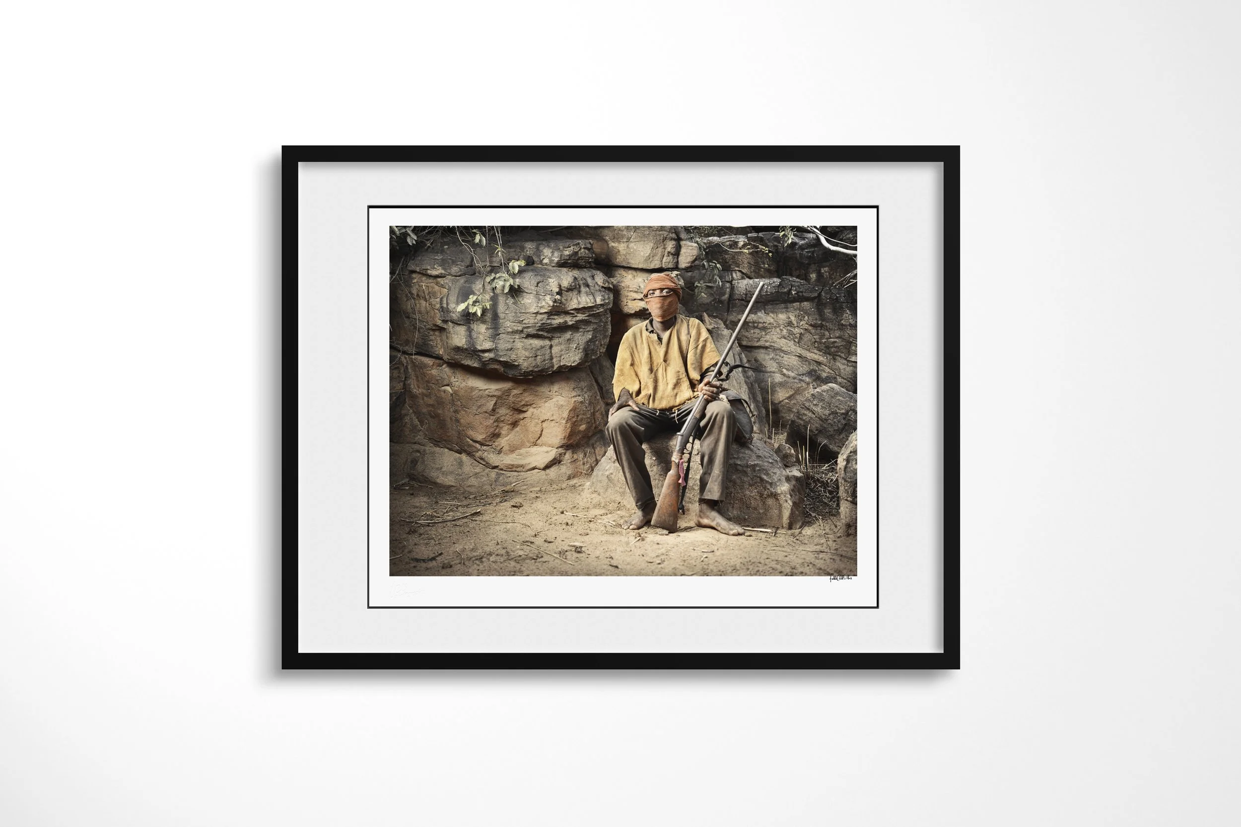 Framed presentation of Duogolou in a black frame and white mount, showing Duogolou seated barefoot against a rock face with his face wrapped and a rifle in hand.
