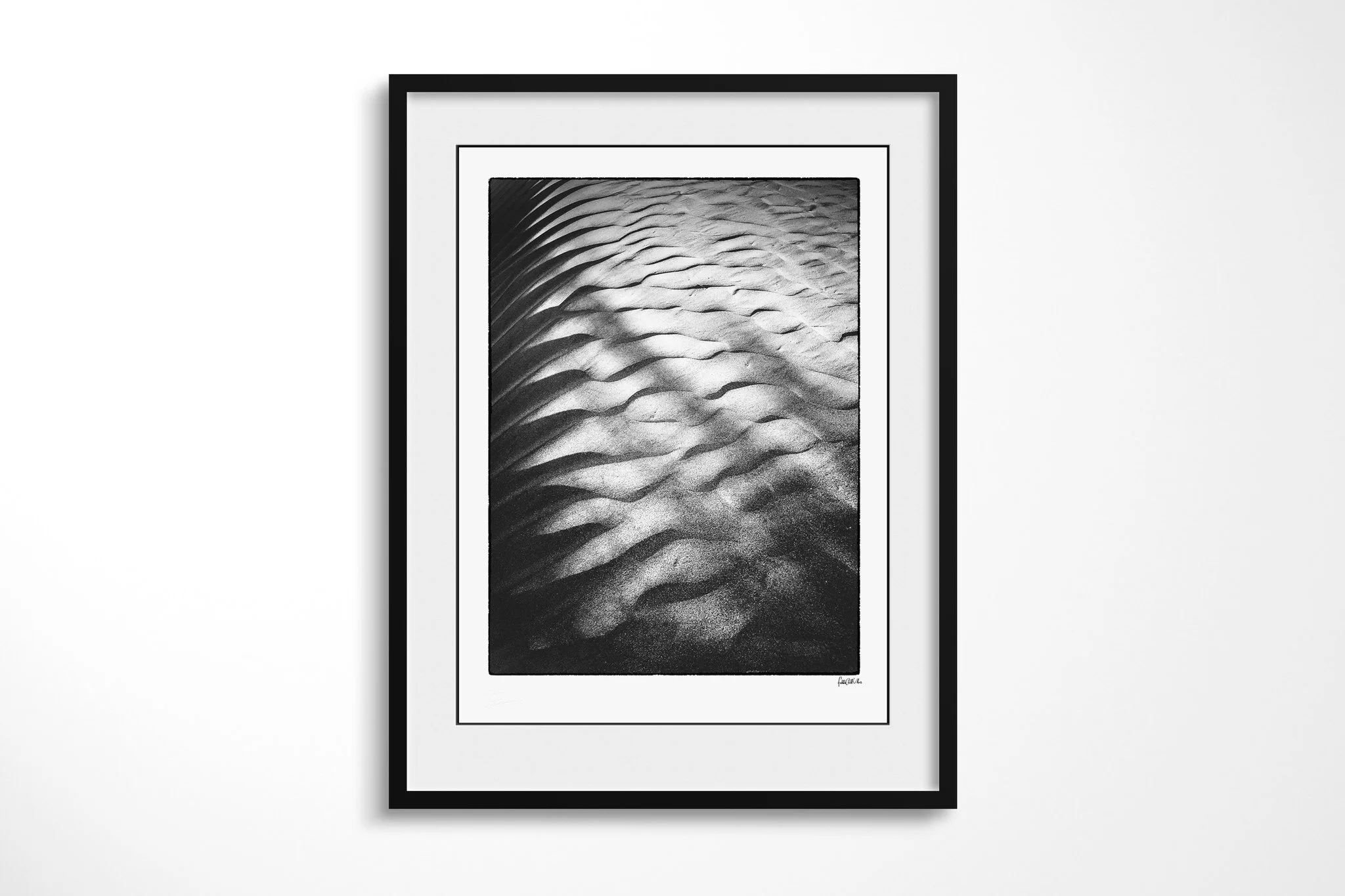 Ripple presented in a black frame and wide white mount, showing a close view of sand ripples with bright crests and deep shadows fading towards the top.