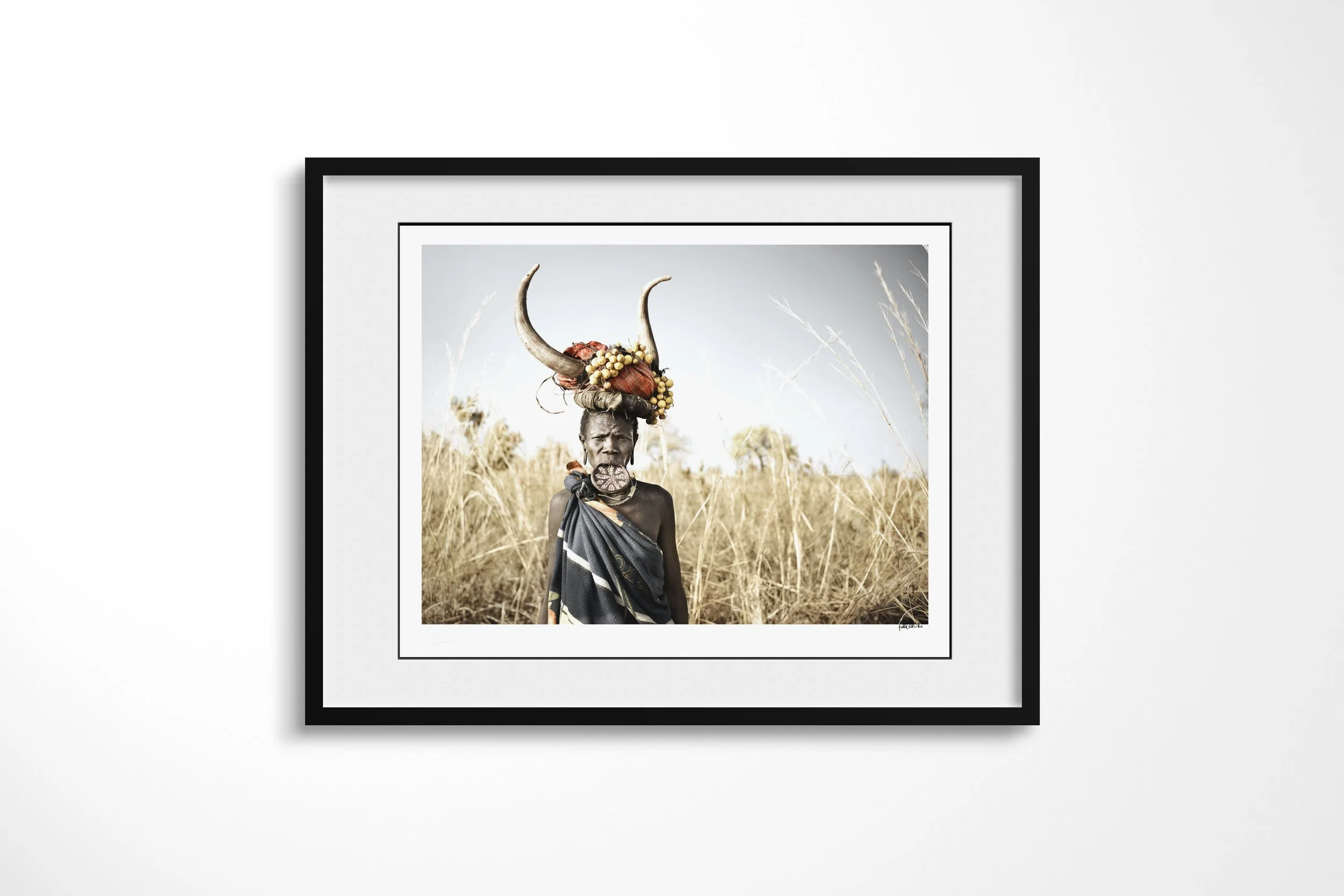 Framed presentation of Makune in a black frame and white mount, showing a Mursi woman wearing a decorated lip plate and curved-horn headdress in tall grass.
