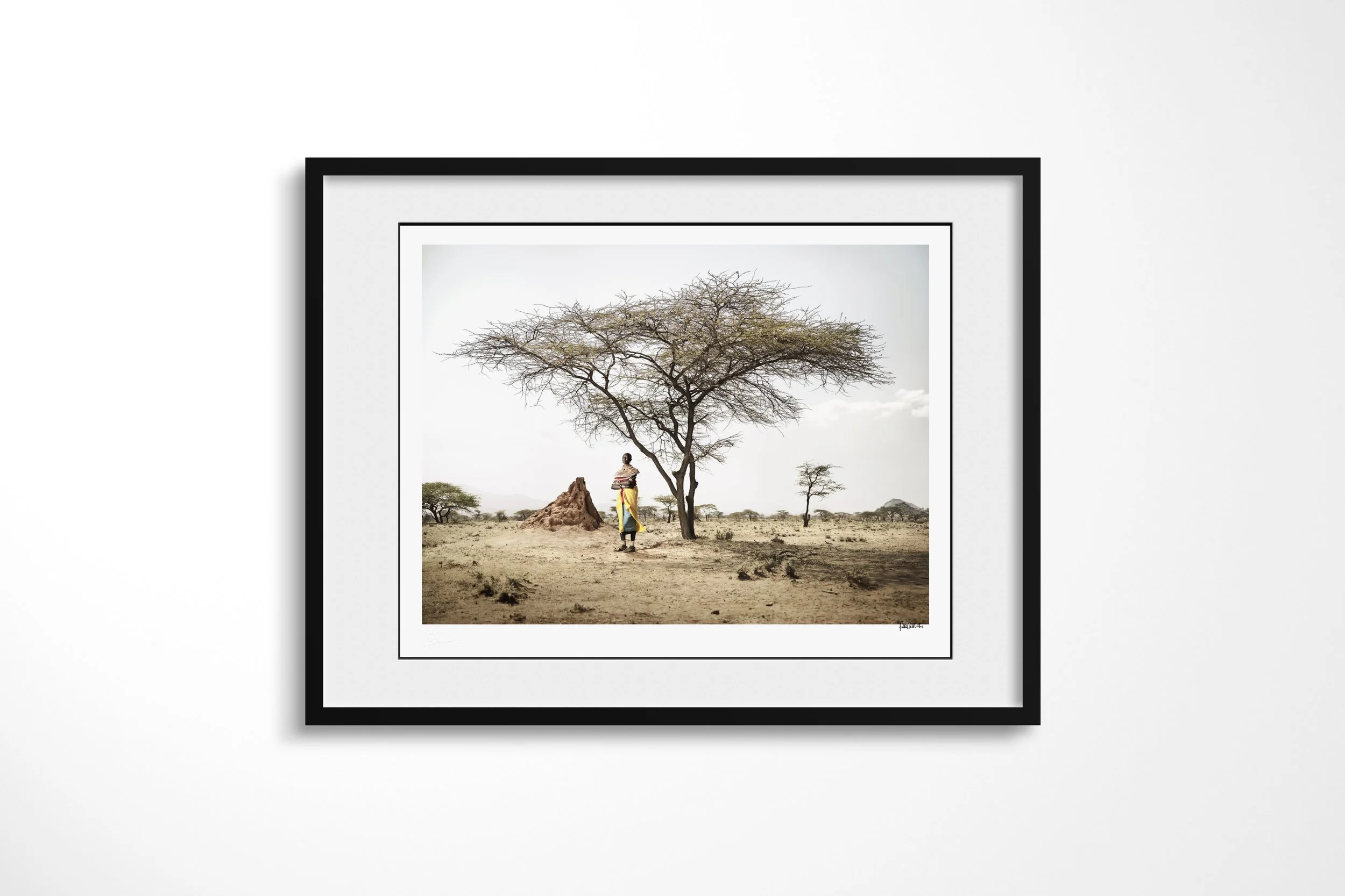 Framed presentation of Kashiumbi in a black frame and white mount, showing a Samburu woman in a bright yellow wrap standing beneath an acacia beside a termite mound.