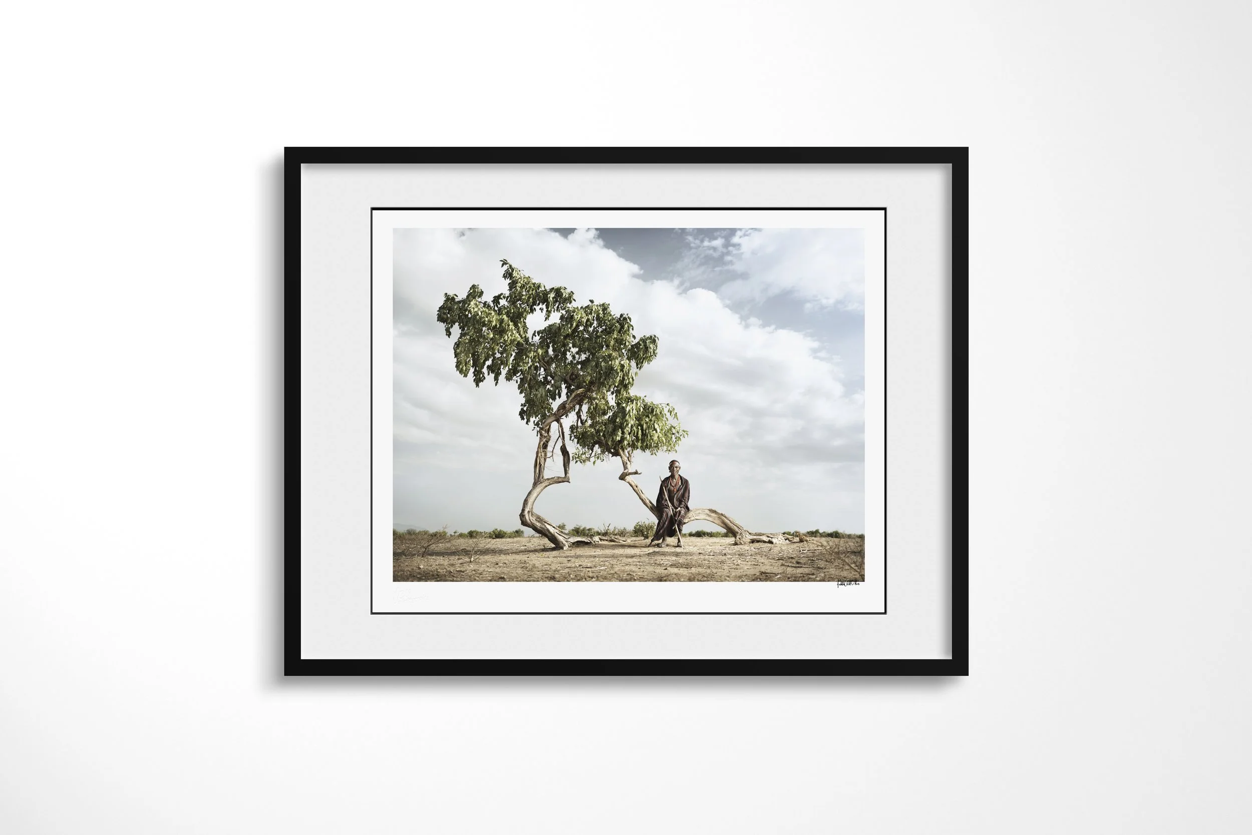 Framed presentation of Gamedu in a black frame and white mount, showing an Arbore man resting beneath the curved trunk of a wind-shaped tree on an open plain.