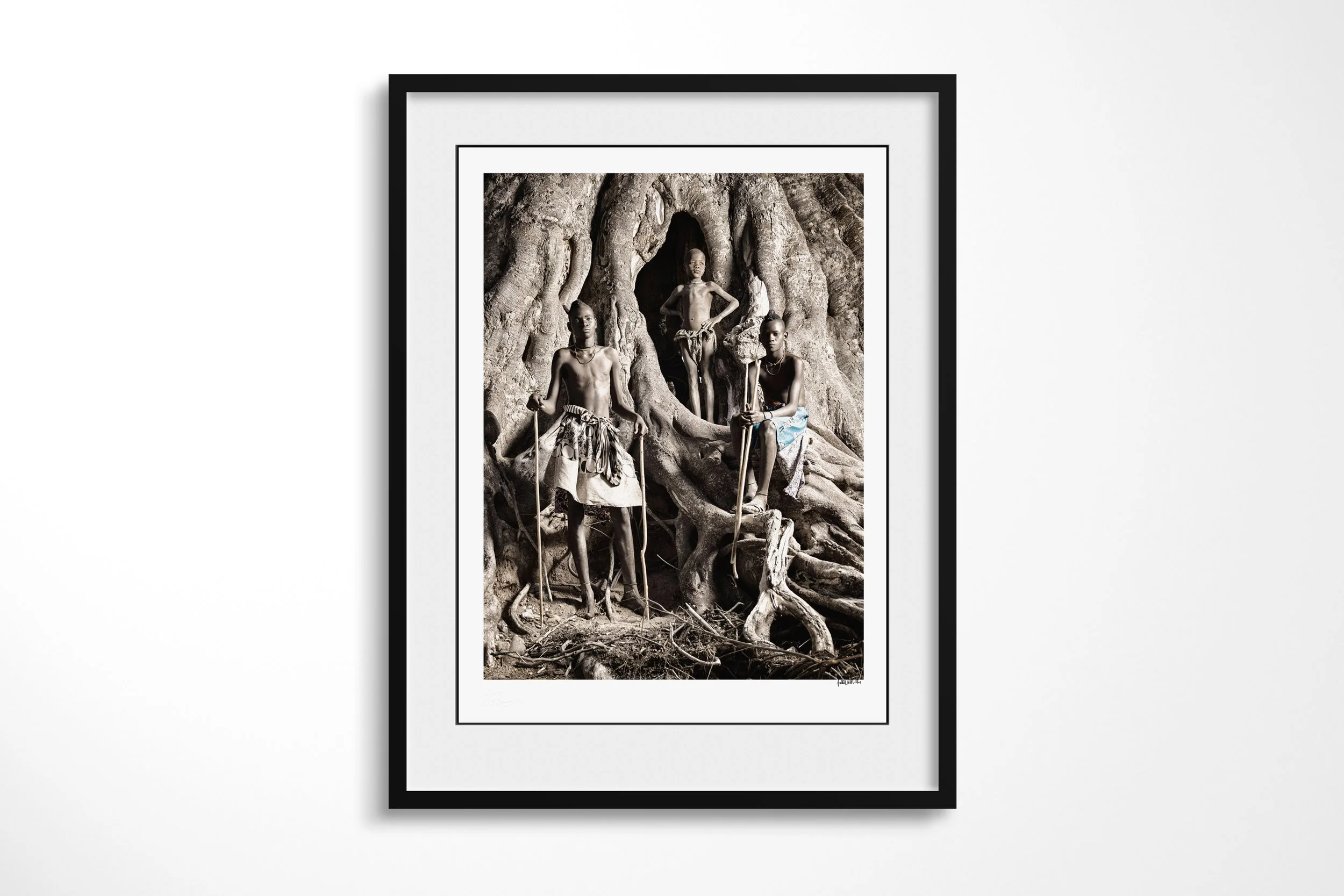 Framed presentation of Turimuno (left), Kakoke (middle) and Mbangu (right) in a black frame and white mount, showing three Himba boys standing among the roots and hollows of a monumental fig tree.