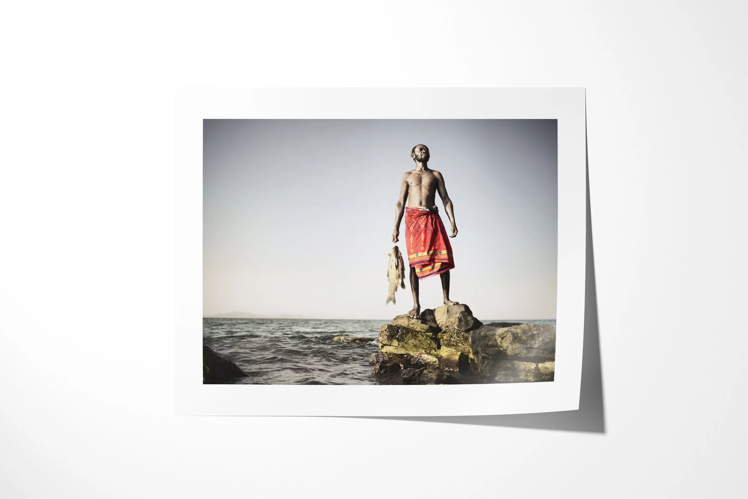 Fine art paper print of Zacheus, showing an El Molo fisherman standing on lakeside rocks holding two fish.