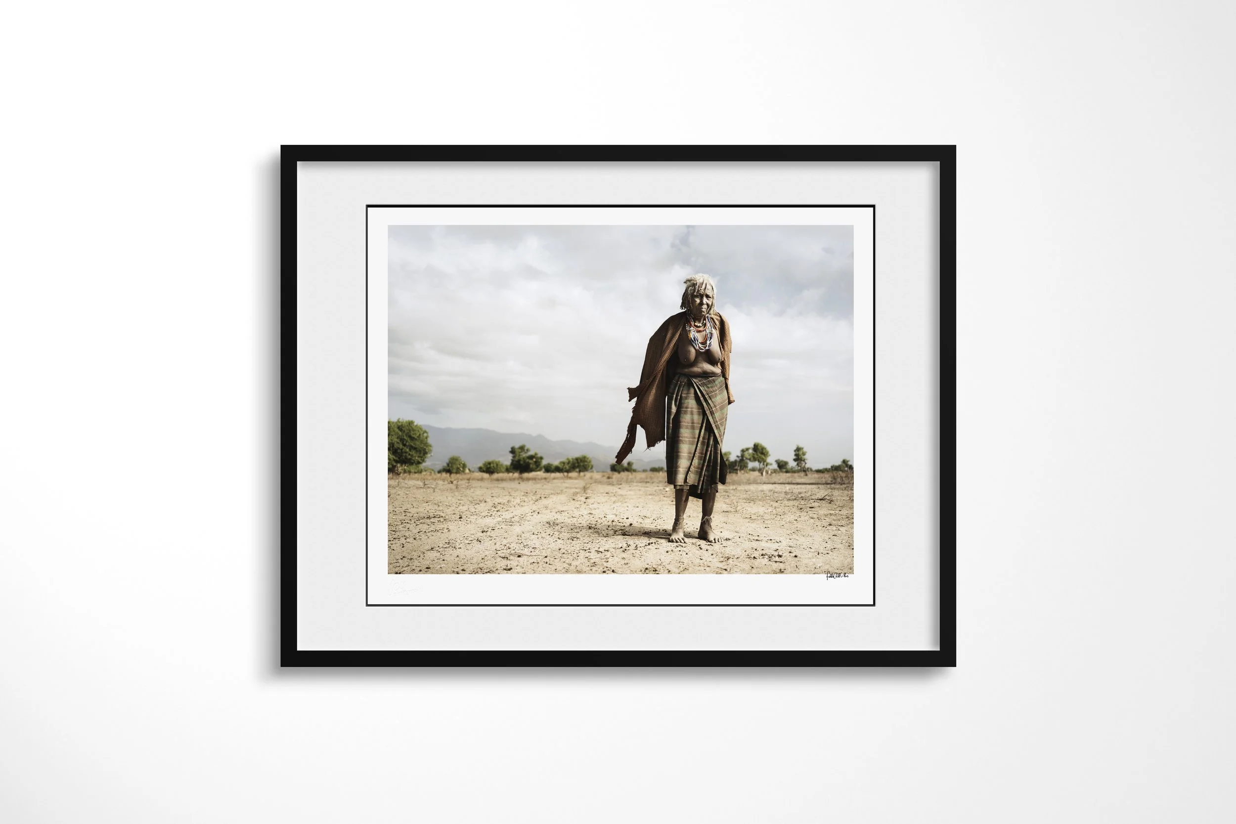 Framed presentation of Rufa in a black frame and white mount, showing an older Arbore woman in layered beadwork and wrapped clothing standing on a dry plain.