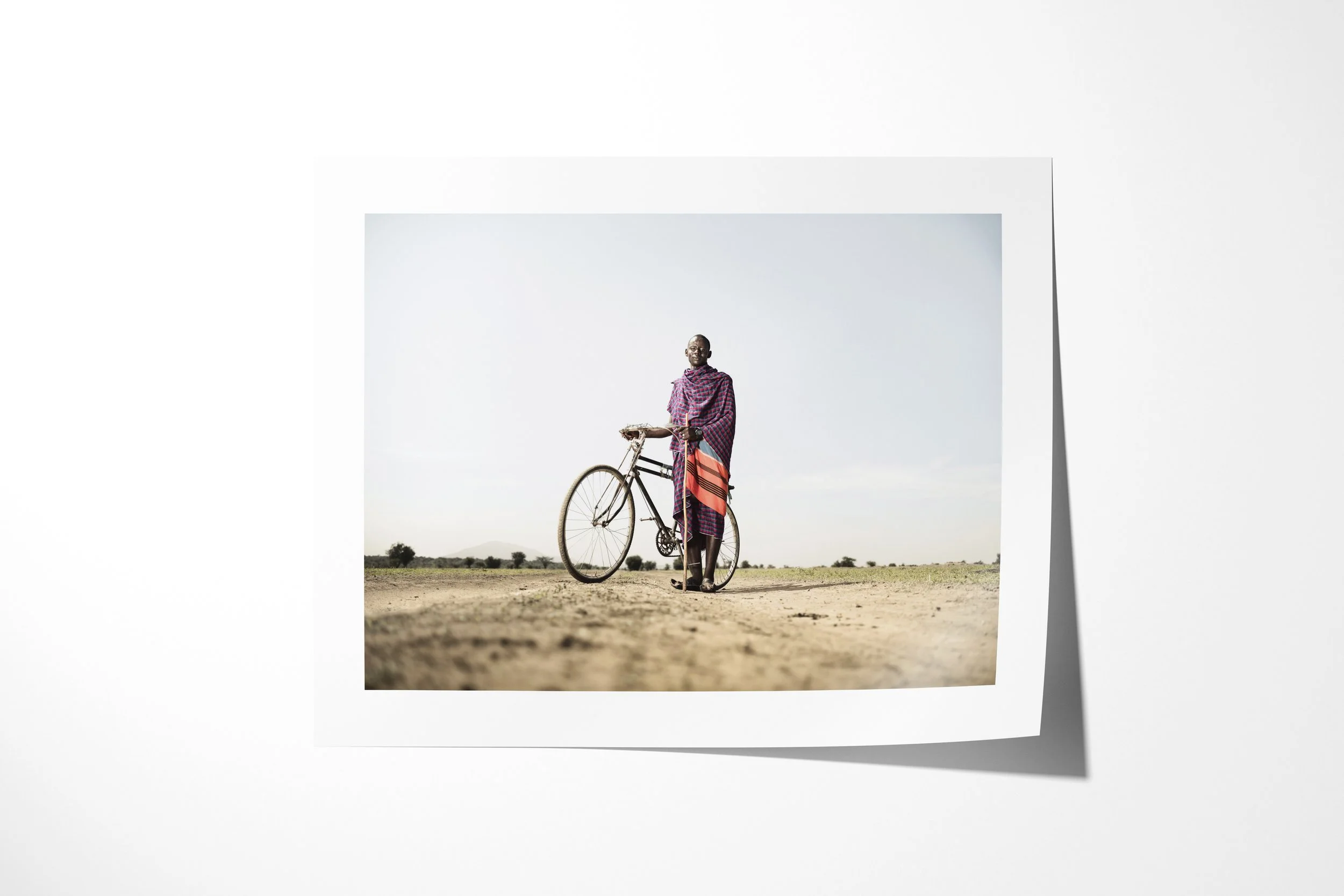 Fine art paper print of Kimerei, showing a Maasai man standing beside his bicycle on an open dirt road.