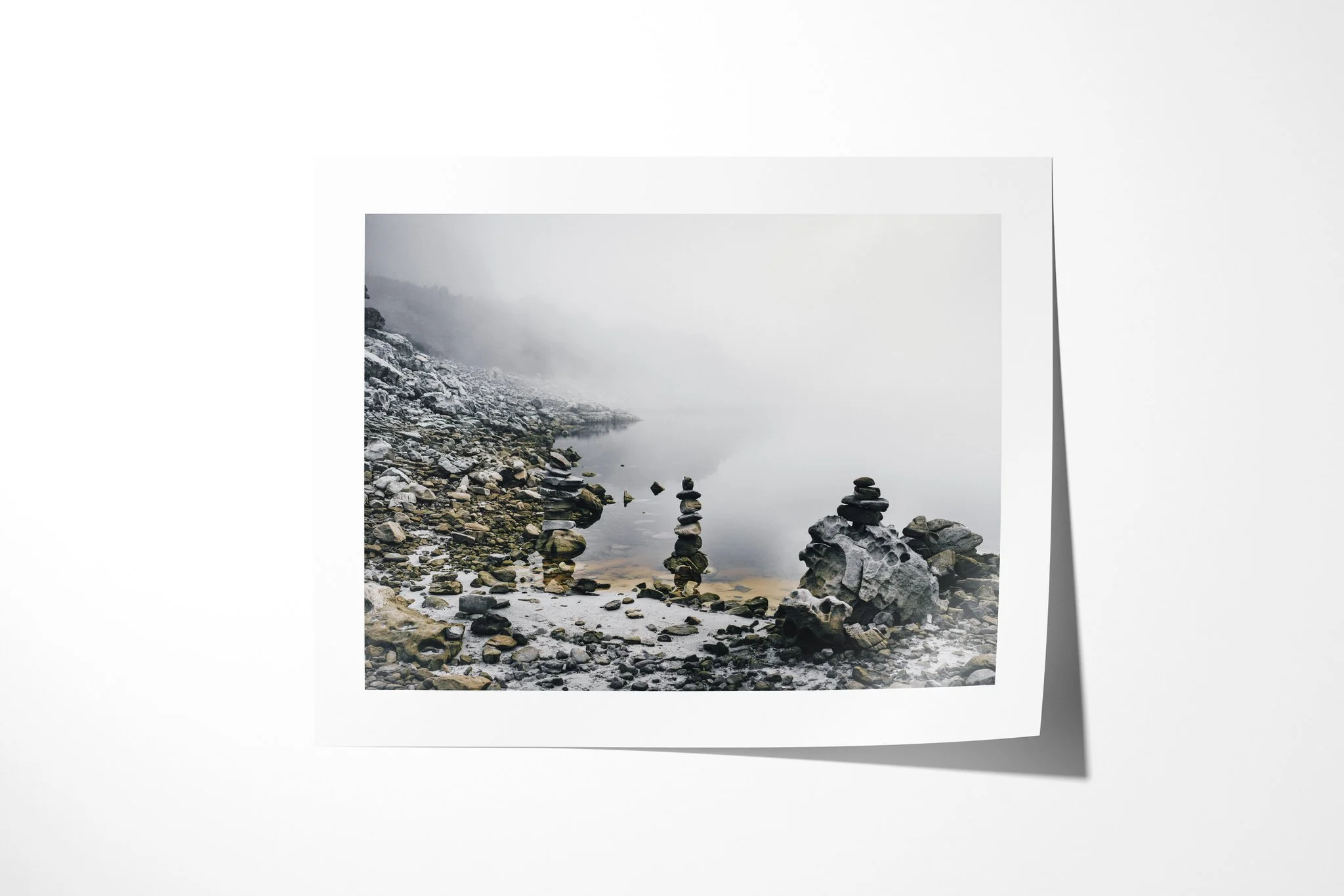 Fine art paper presentation of Cairn, depicting balanced stone cairns beside still water on a rocky shore fading into pale fog.