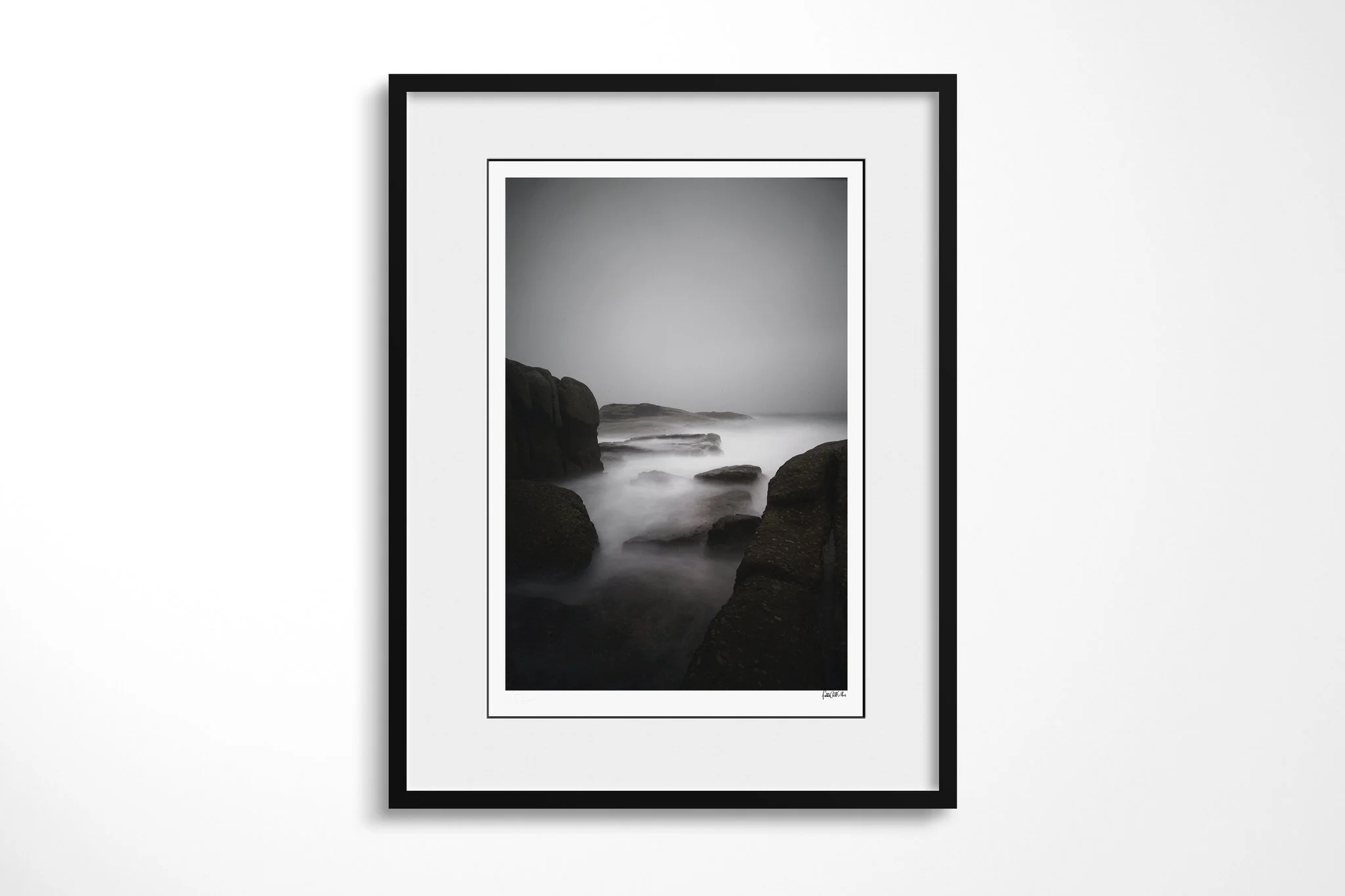 Framed presentation of Misty Morning in a black frame and white mount, showing dark coastal boulders framing pale long-exposure seawater beneath a blank grey mist.
