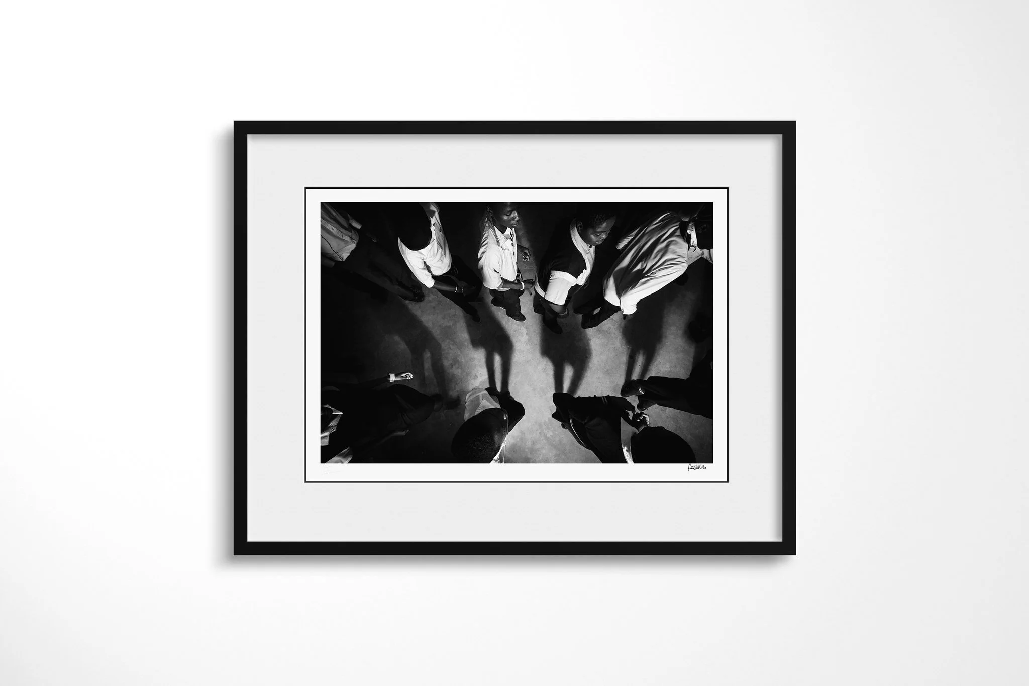 Black-framed fine art print of Chant with a white mount, showing a group of people seen from directly overhead, standing in a loose circle with long shadows across a concrete floor.