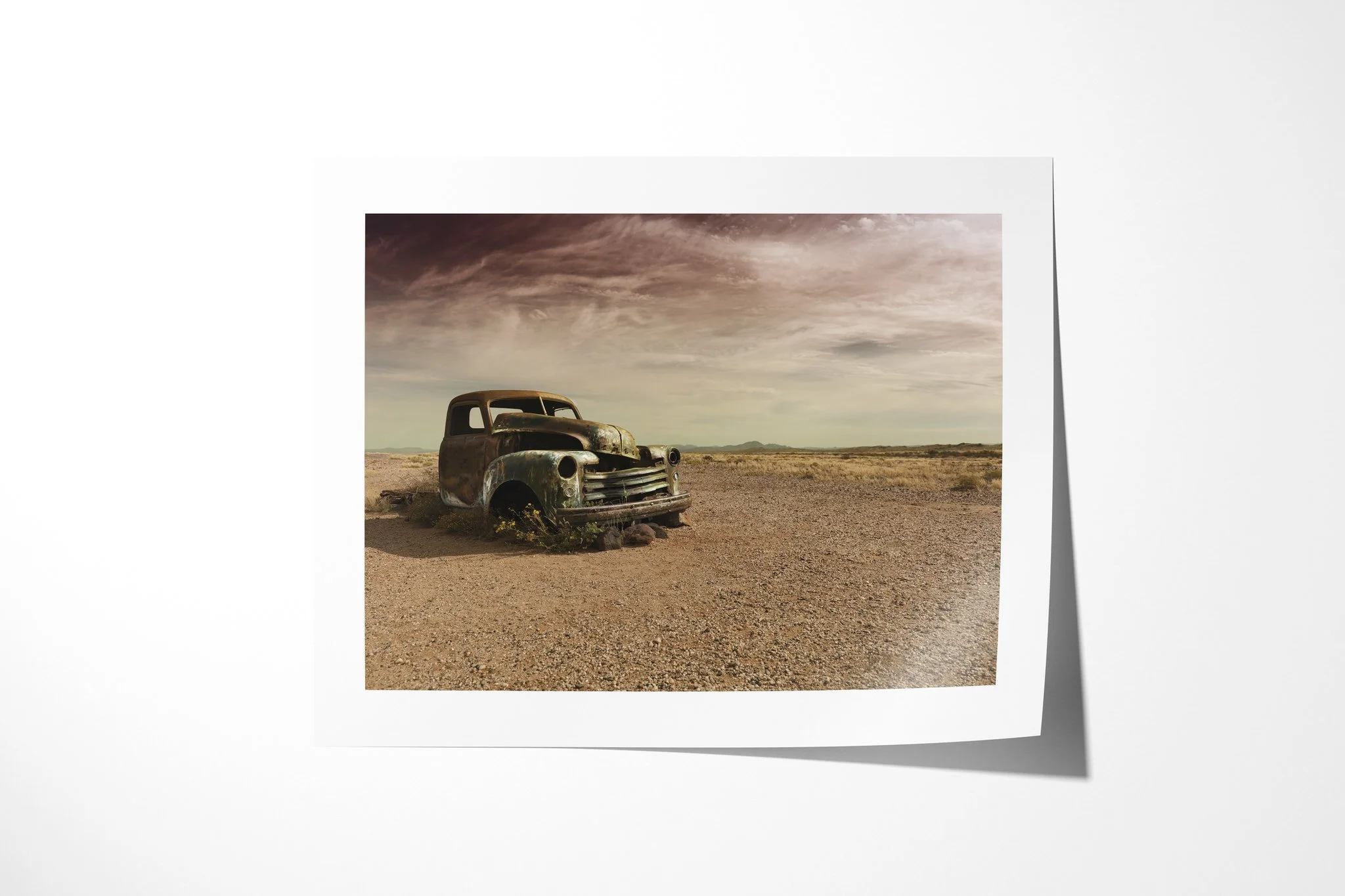 Fine art paper print of Better Days, showing a rusted vintage vehicle abandoned on a stony arid plain beneath dark, textured clouds and distant low mountains.