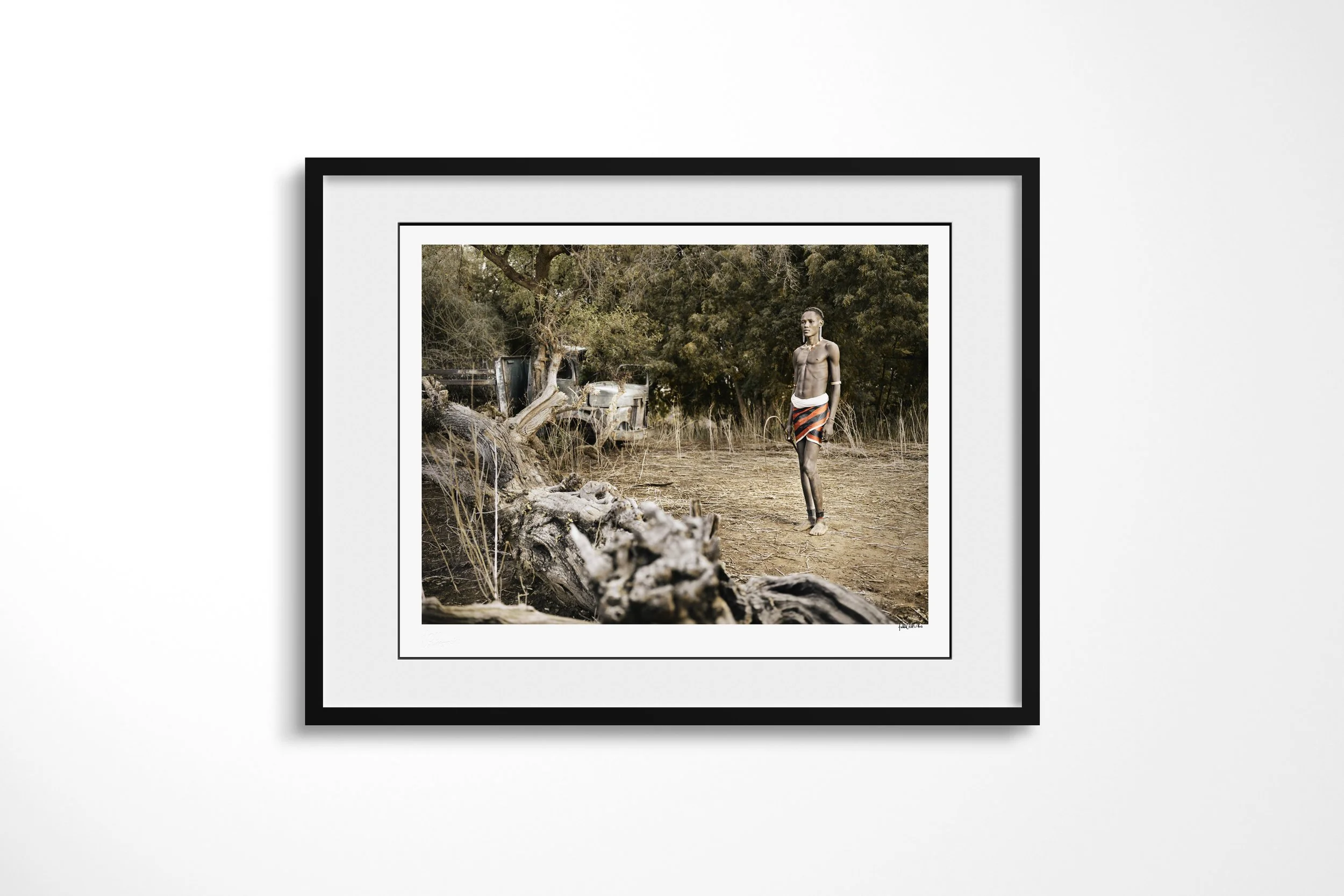 Framed presentation of Lokoloi in a black frame and white mount, showing a Daasanach man standing near an uprooted tree with an abandoned vehicle behind him.