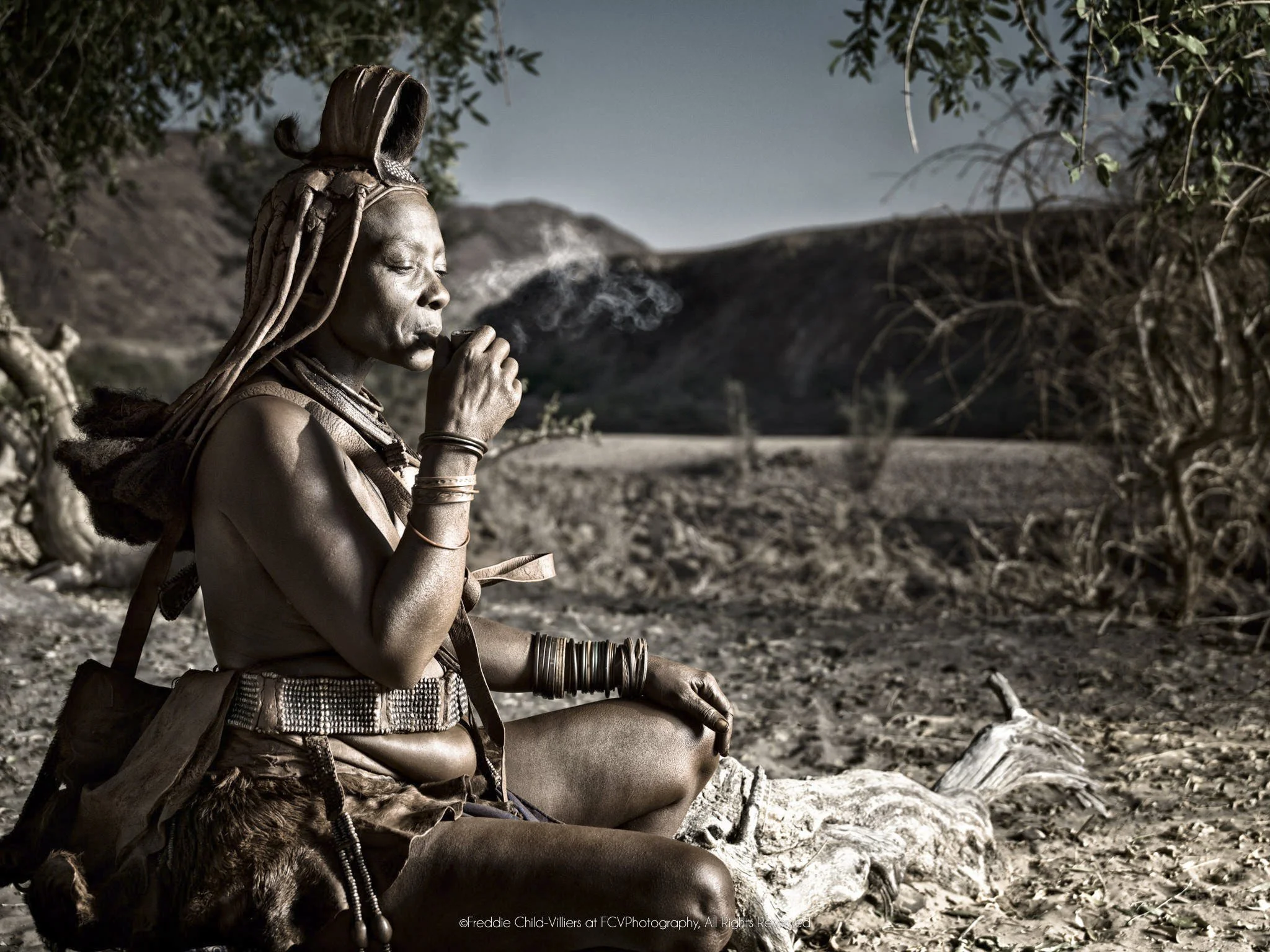 Ngorera seated beneath a tree smoking a small pipe in the dry Kunene landscape.