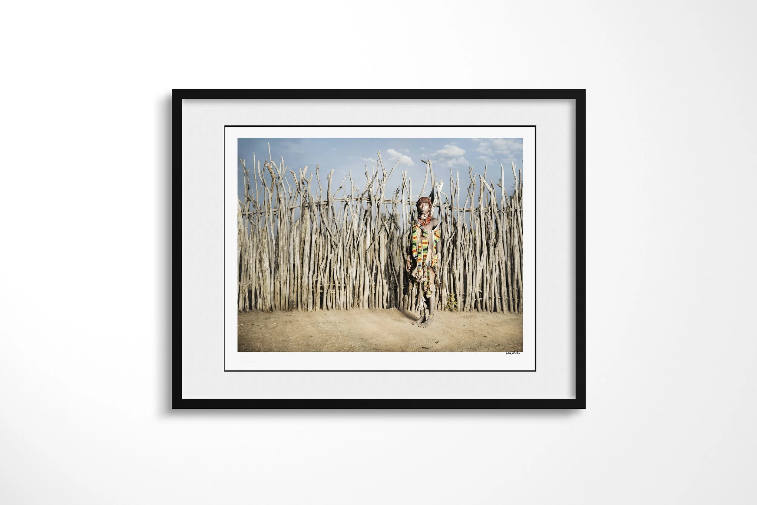 Framed presentation of Aito in a black frame and white mount, showing a Hamar woman in layered beadwork and decorated hide clothing standing against a tall wooden fence.